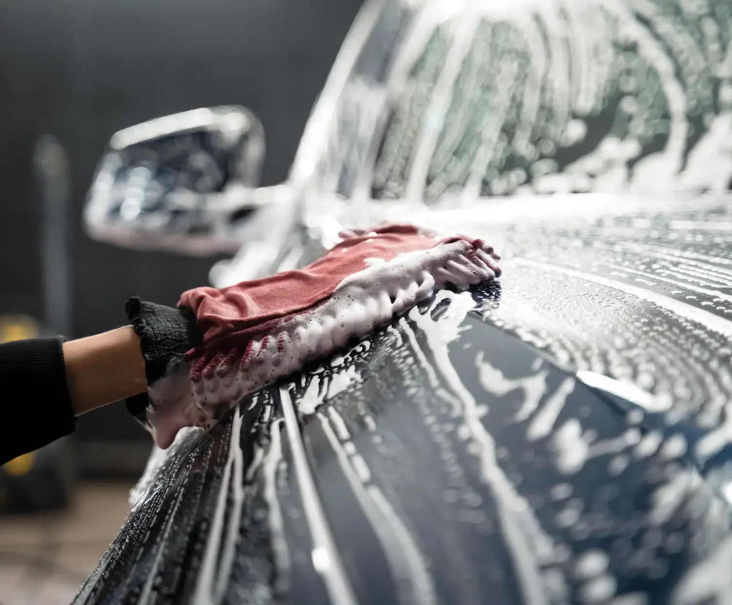 A person using a red sponge to wash a car, with soap suds on the surface.