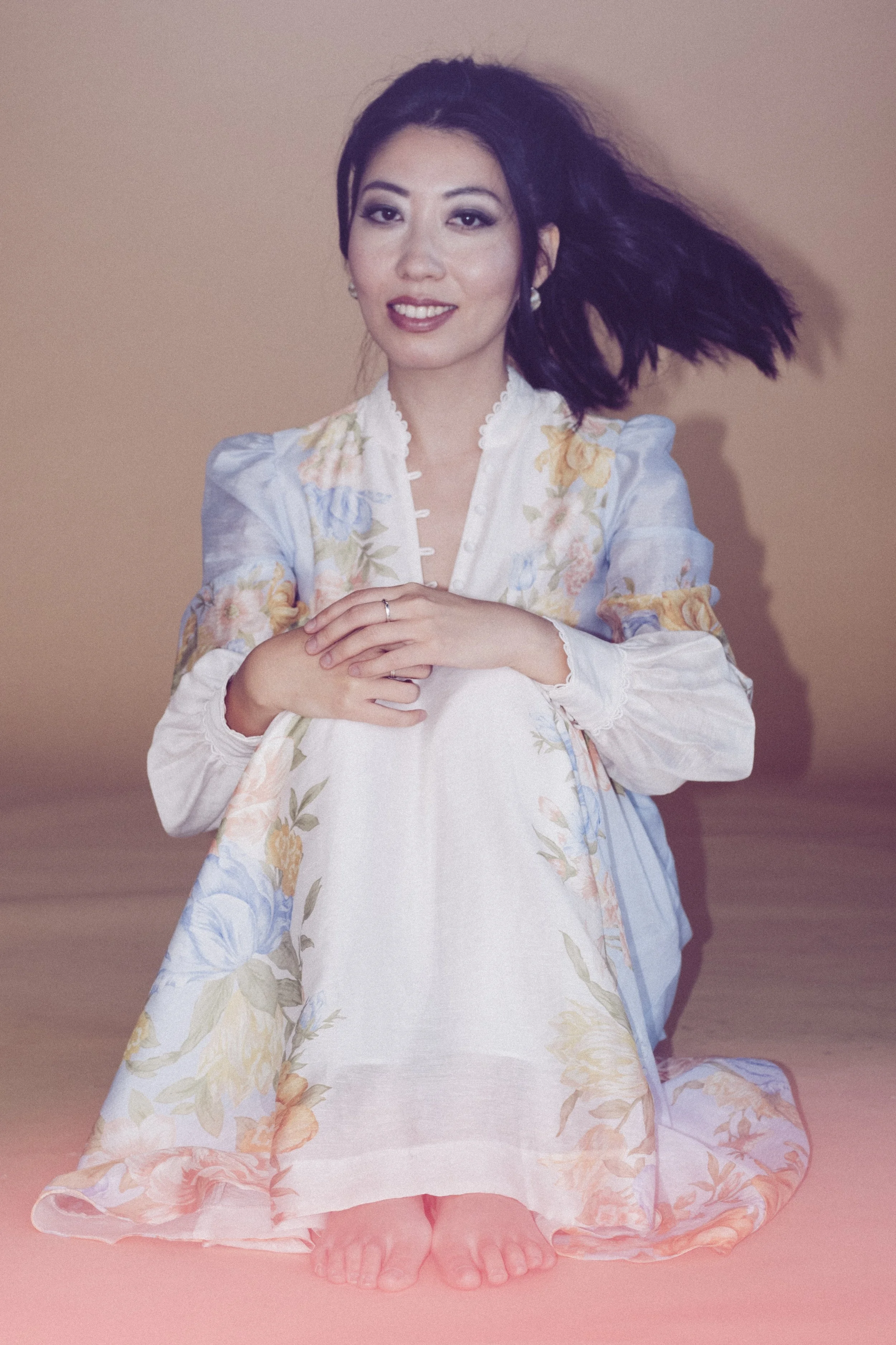 Wanting Qu wearing a floral dress and earrings, kneeling on the floor with her hands clasped in front of her, smiling, against a neutral background.