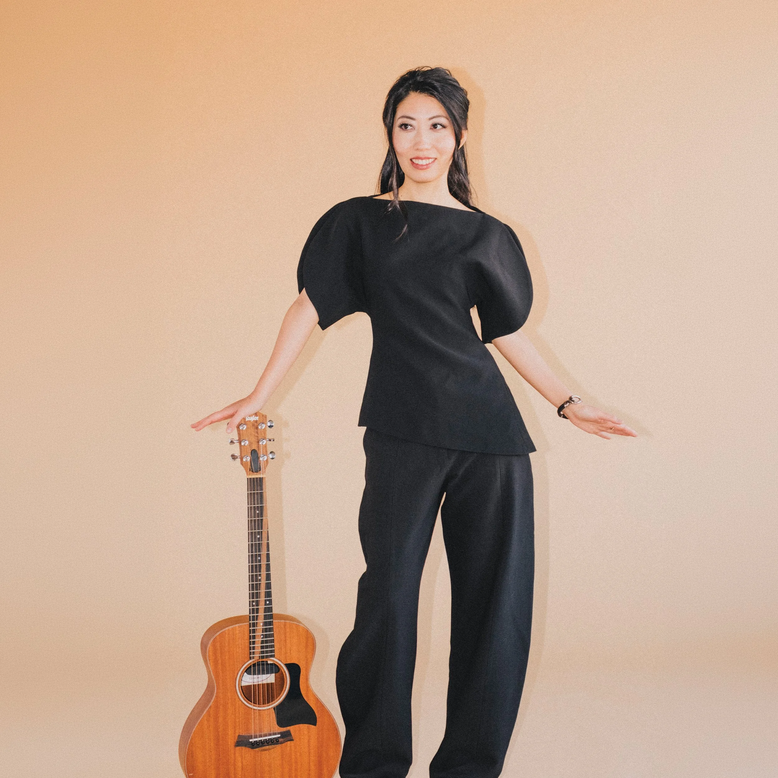 Wanting Qu wearing a black dress posing next to a wooden acoustic guitar against a beige background.