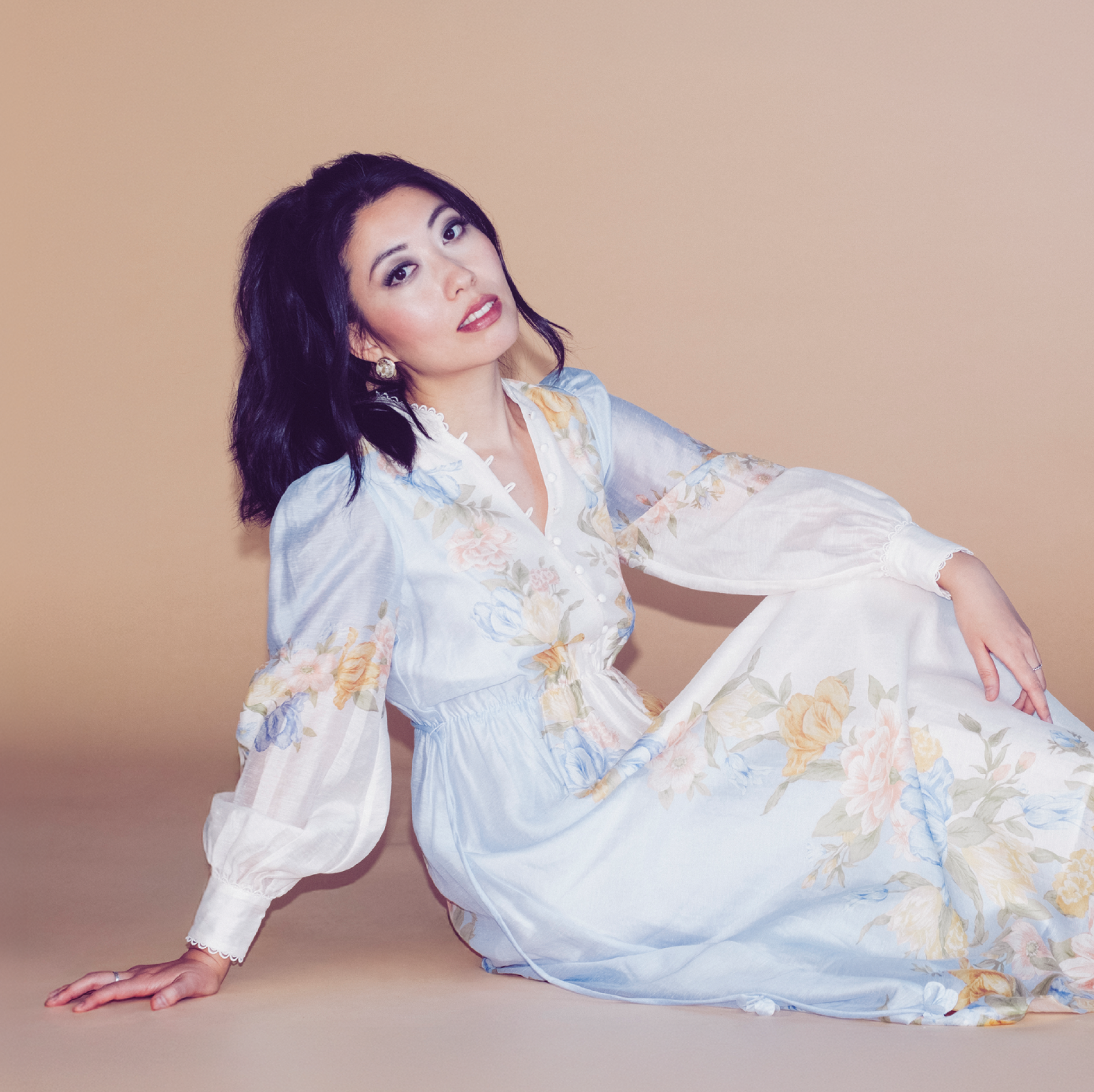Wanting Qu wearing a pastel floral dress, is sitting on the floor against a plain beige background. She is leaning on her left arm with her hand on the ground, looking into the camera with a slight smile.