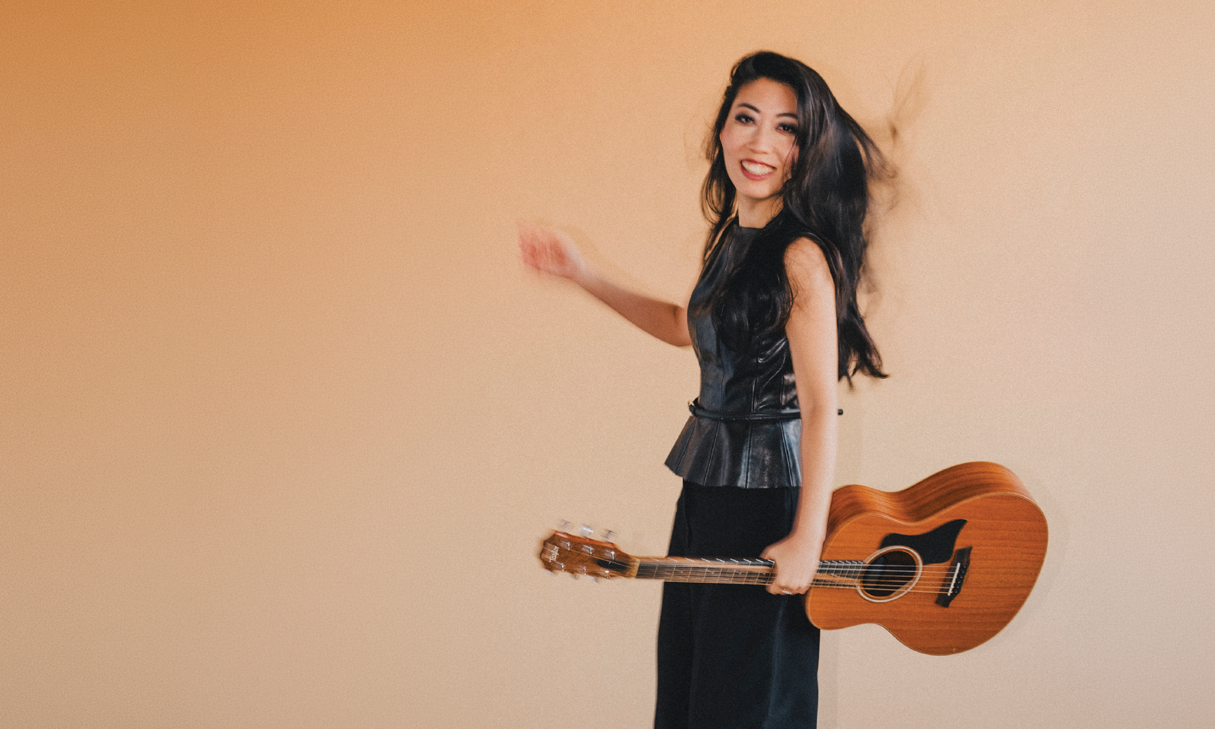Wanting Qu smiling and looking at the camera, wearing a black leather top, standing against a plain beige wall, holding an acoustic guitar.