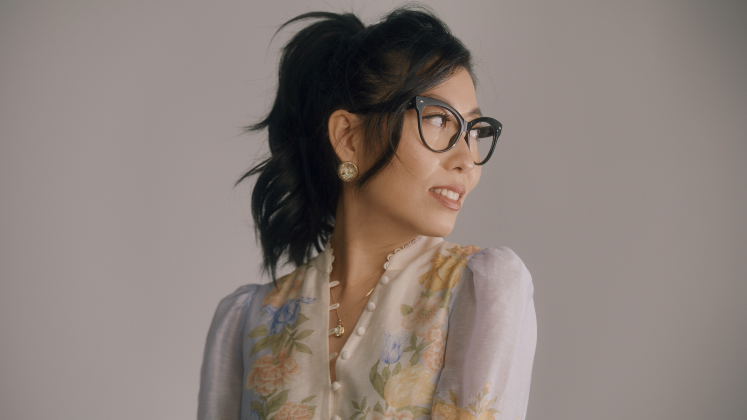 Wanting Qu wearing glasses, gold earrings, and a floral dress, looking to her left against a plain light grey background.