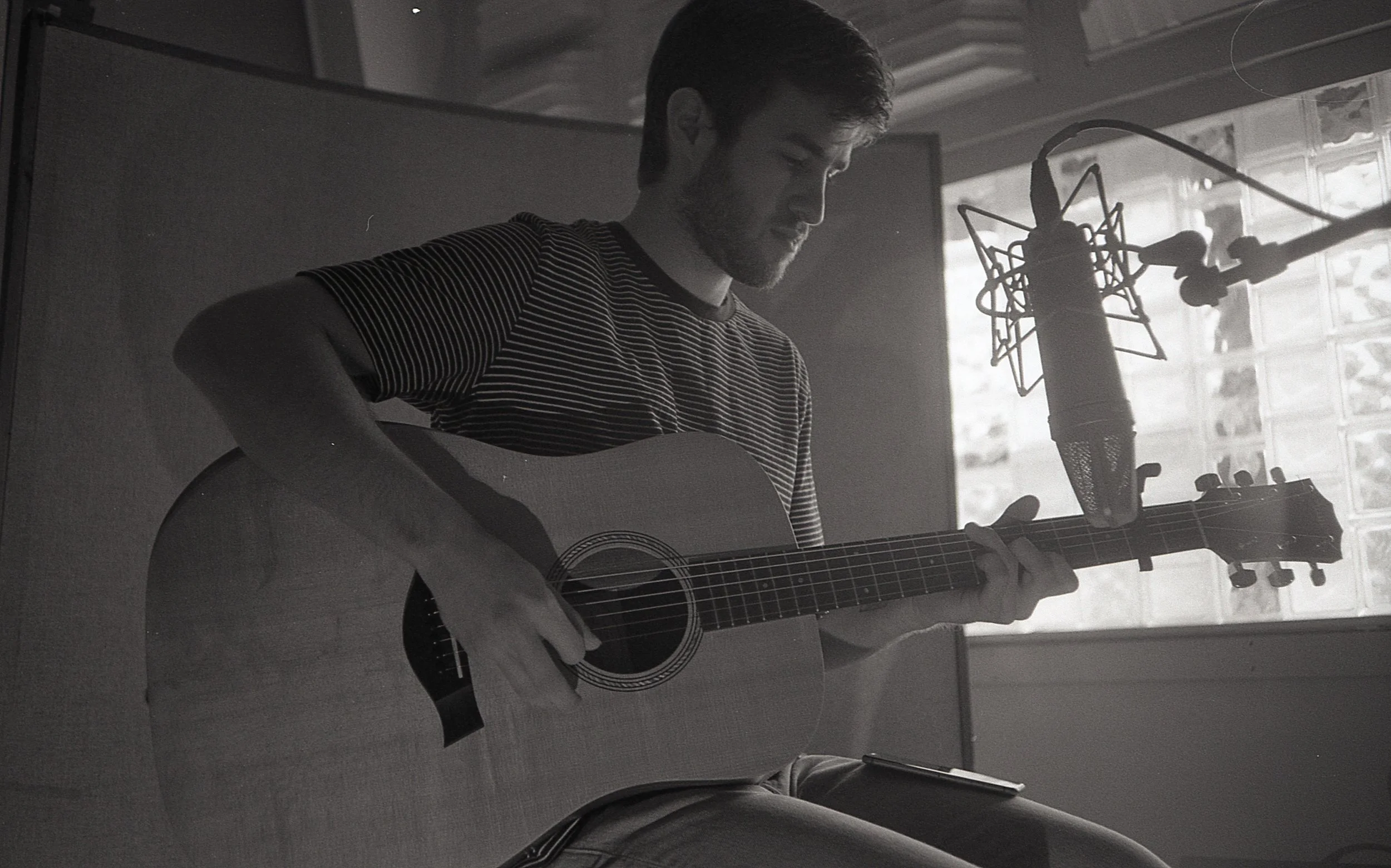 A man with dark hair and a beard playing an acoustic guitar in a recording studio, positioned near a large microphone with a pop filter, with a background of a window letting in natural light.