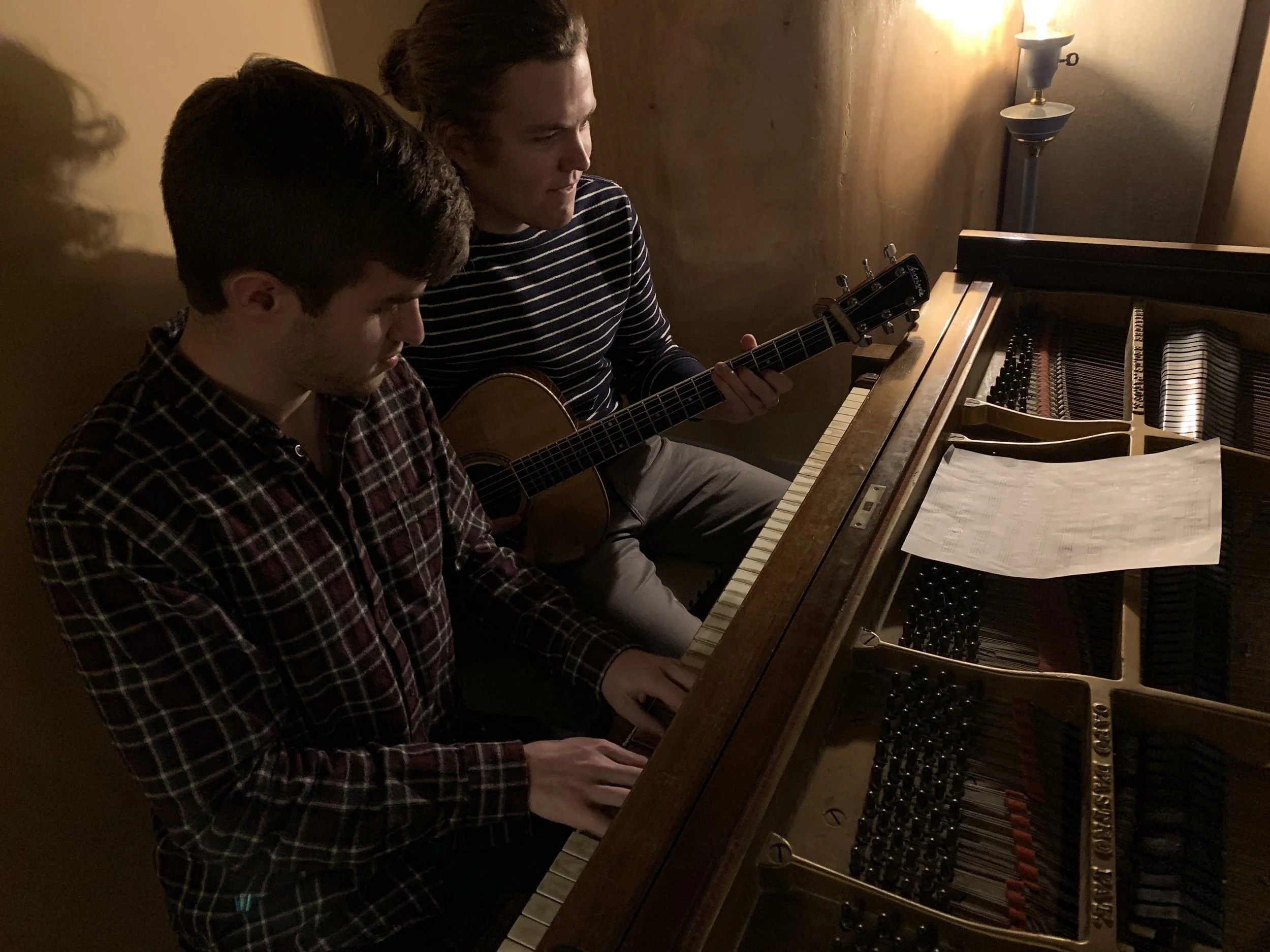 Two young men playing a piano and guitar together in a recording studio with ambient lighting.