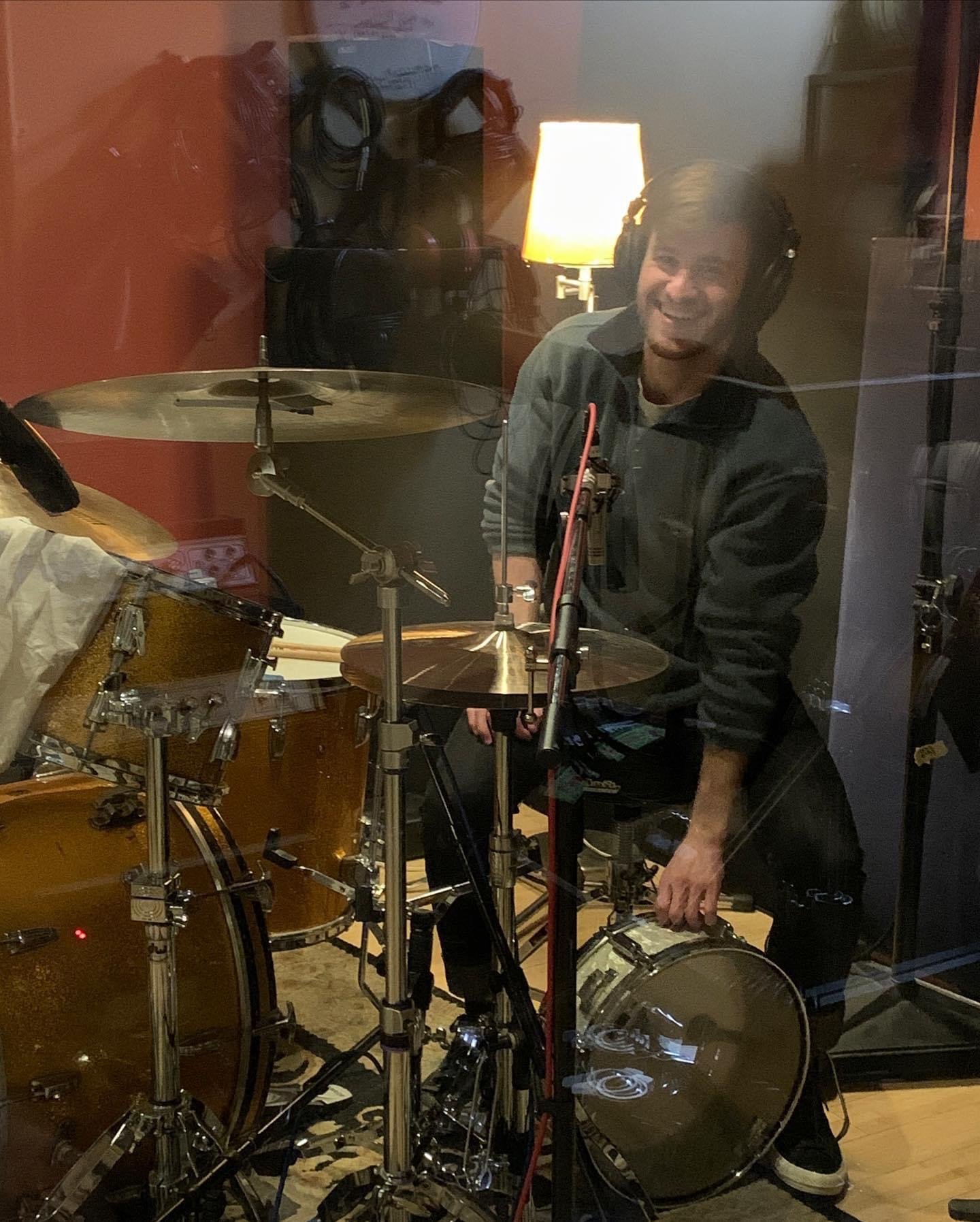 A musician in a recording studio, smiling at the camera, standing behind a drum set and wearing headphones, with a lamp and other recording equipment visible in the background.