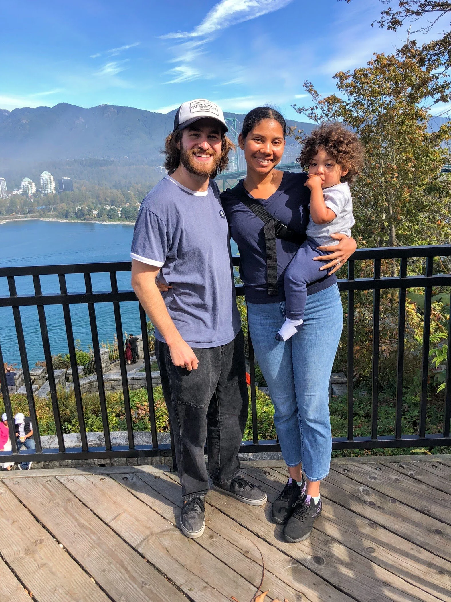 Three people standing on a wooden deck with a scenic background of water, mountains, and trees. One man, woman, and a young child. The woman is holding the child. All are smiling.