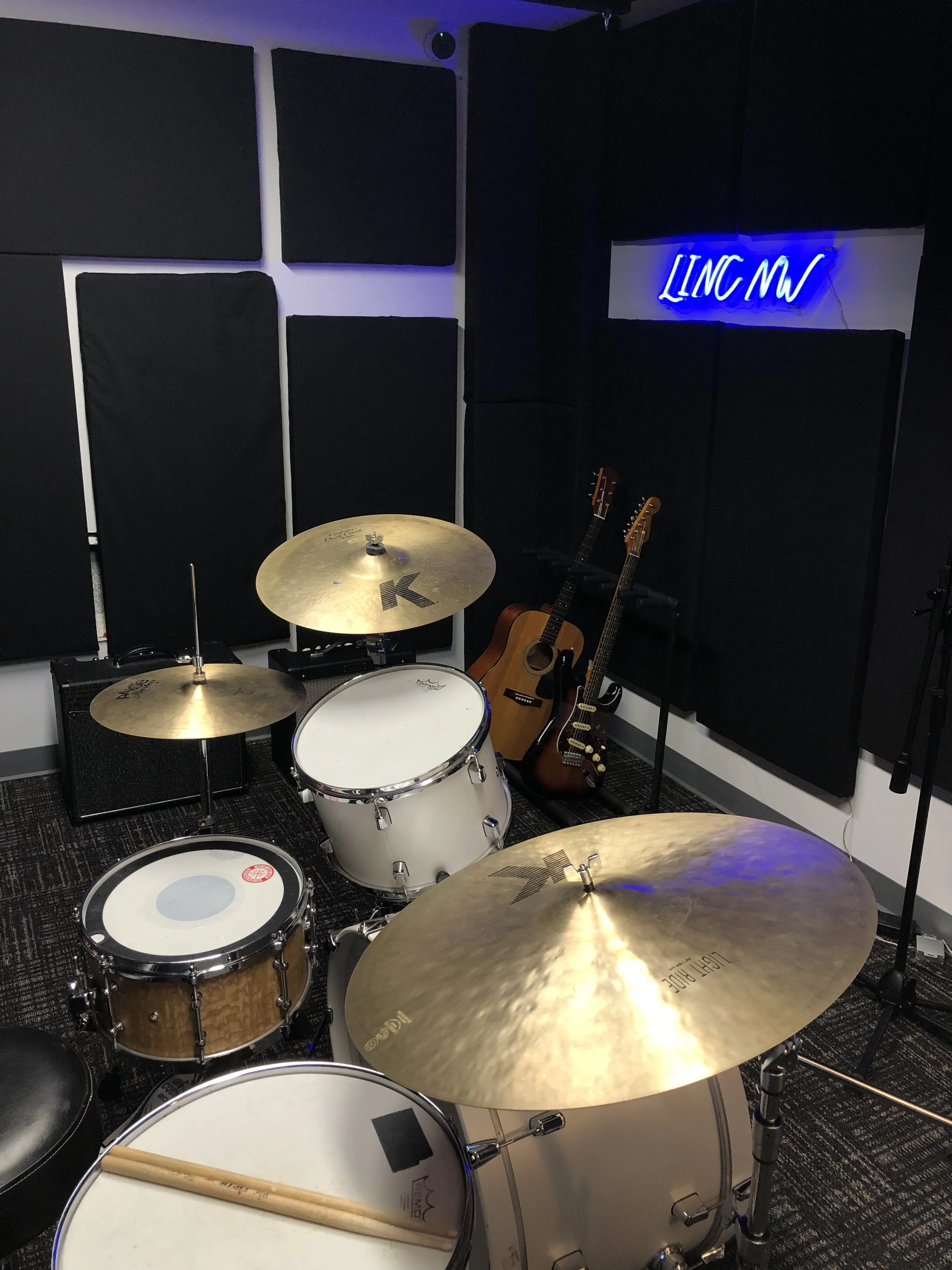 A music rehearsal room with two acoustic guitars, a bass guitar, and a drum set, illuminated by a blue neon sign that reads "LINC MI" on black acoustic foam-covered walls.