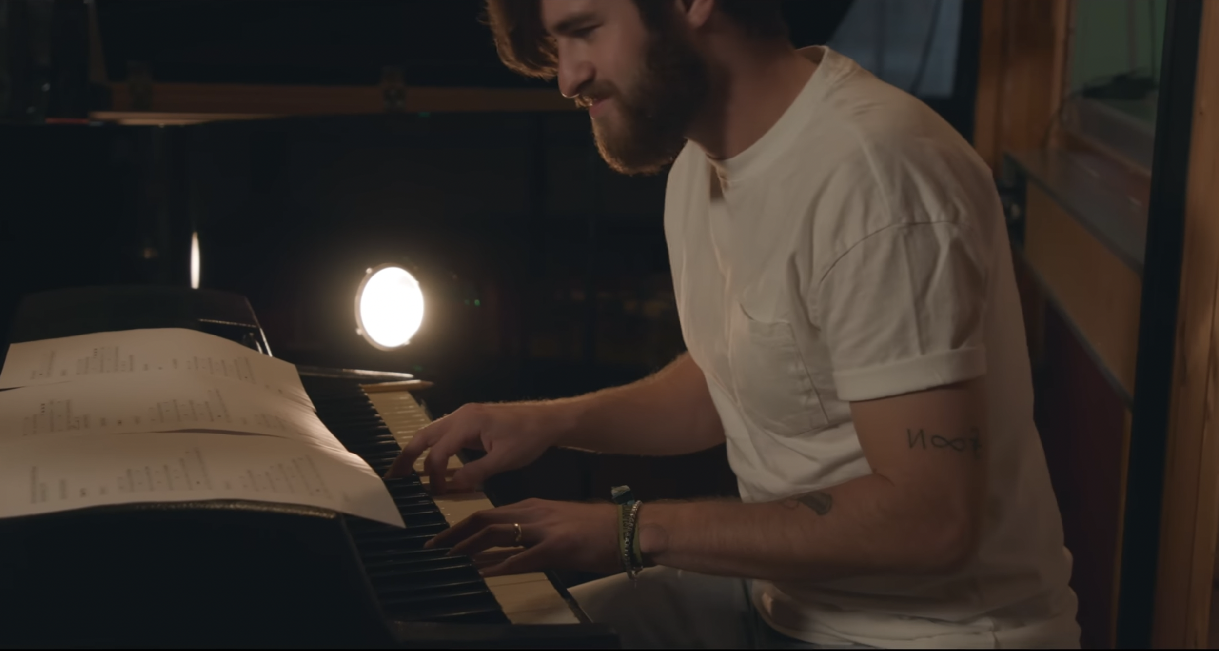 A man with a beard and tattoos wearing a white t-shirt is playing a keyboard with sheet music on top in a dimly lit room.
