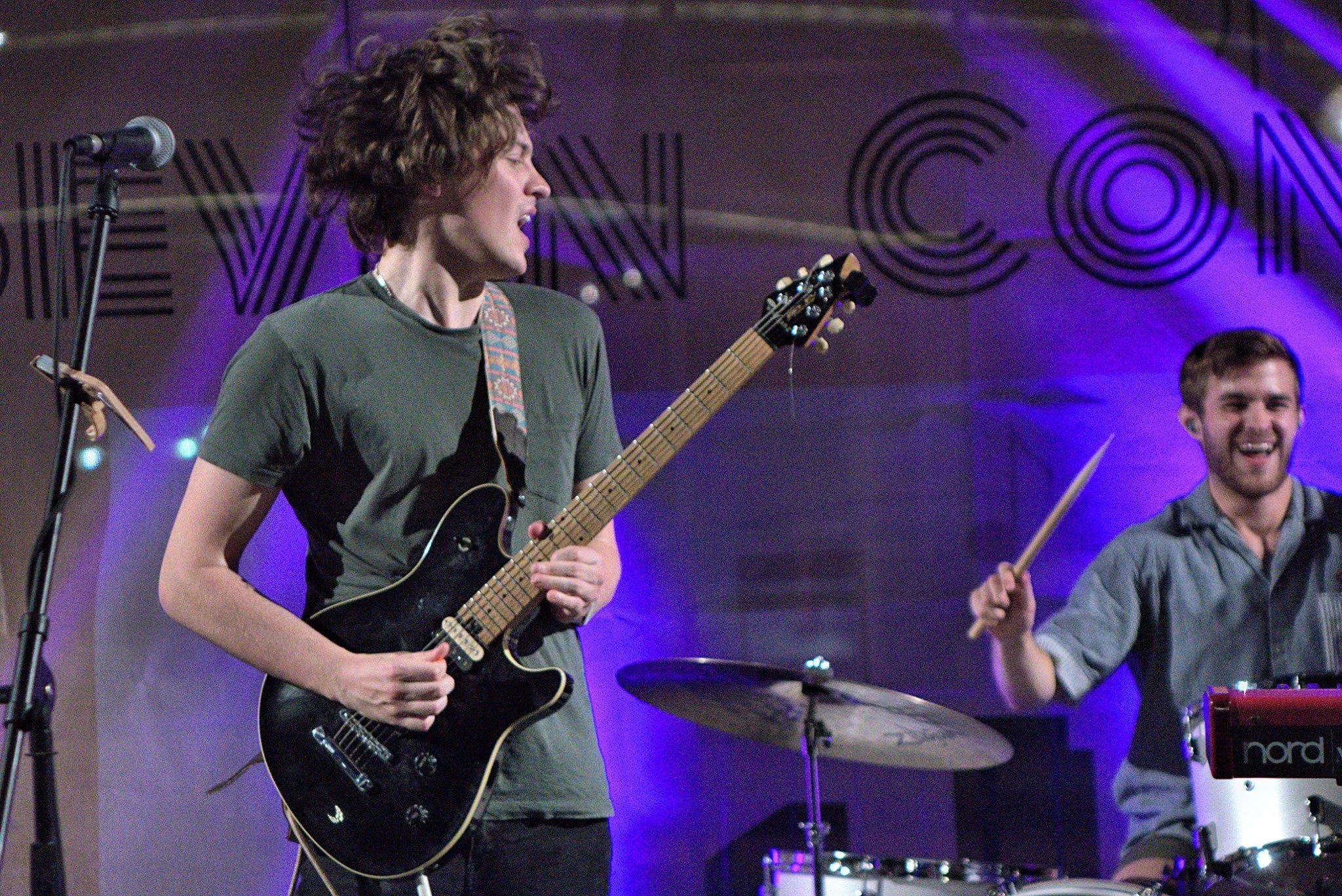 Two musicians performing on stage in Downtown Boston. One playing an electric guitar, the other on drums, both smiling and engaged with their instruments.