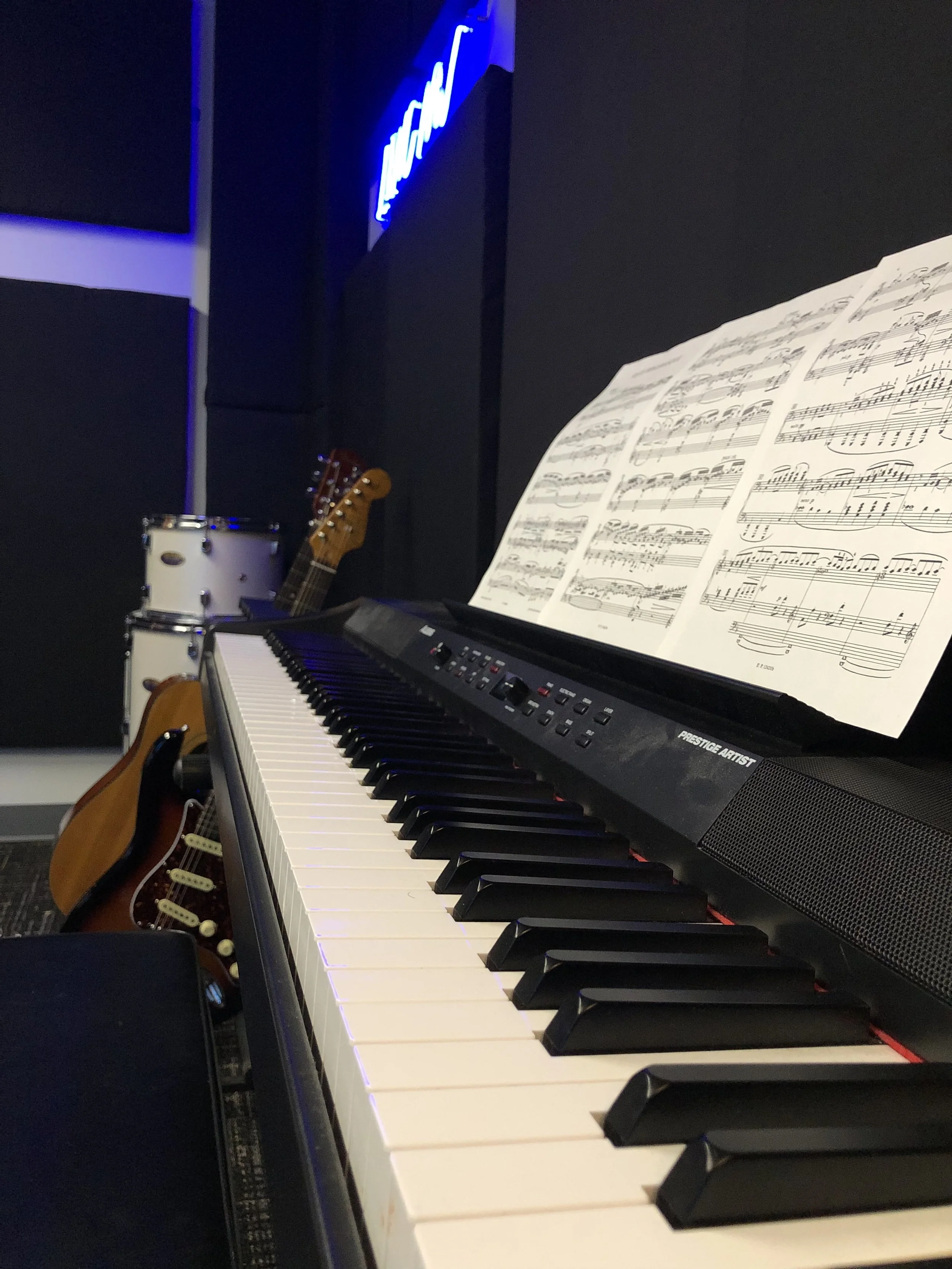 An electronic keyboard with sheet music on display, a guitar in a stand next to a stack of drums, and a neon sign in the background in a music studio.