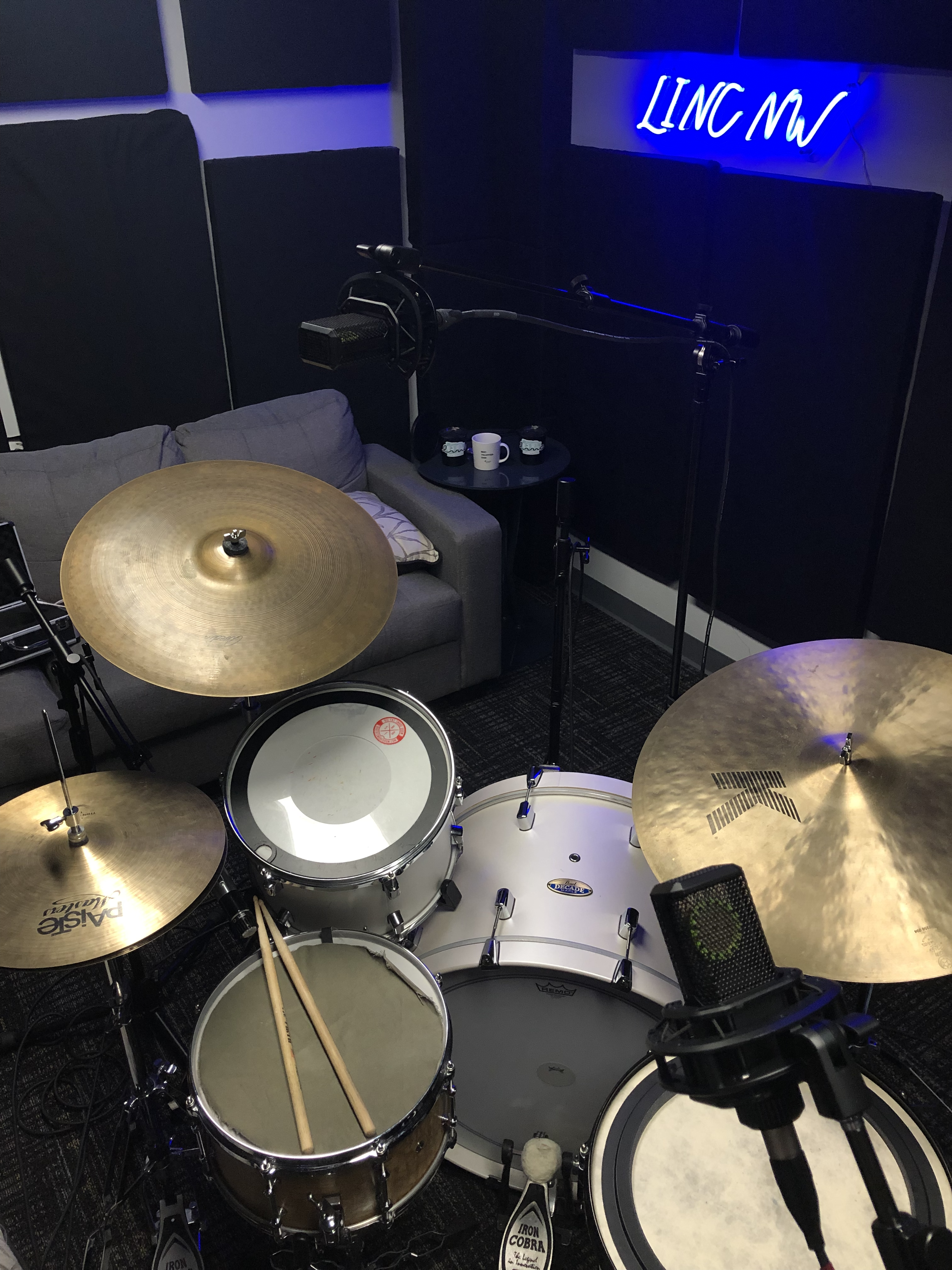 Music recording studio with drums and cymbals, a microphone on a stand, a sofa, coffee cups, and a neon sign that says 'LINC NW' in a dark room with black acoustic panels.