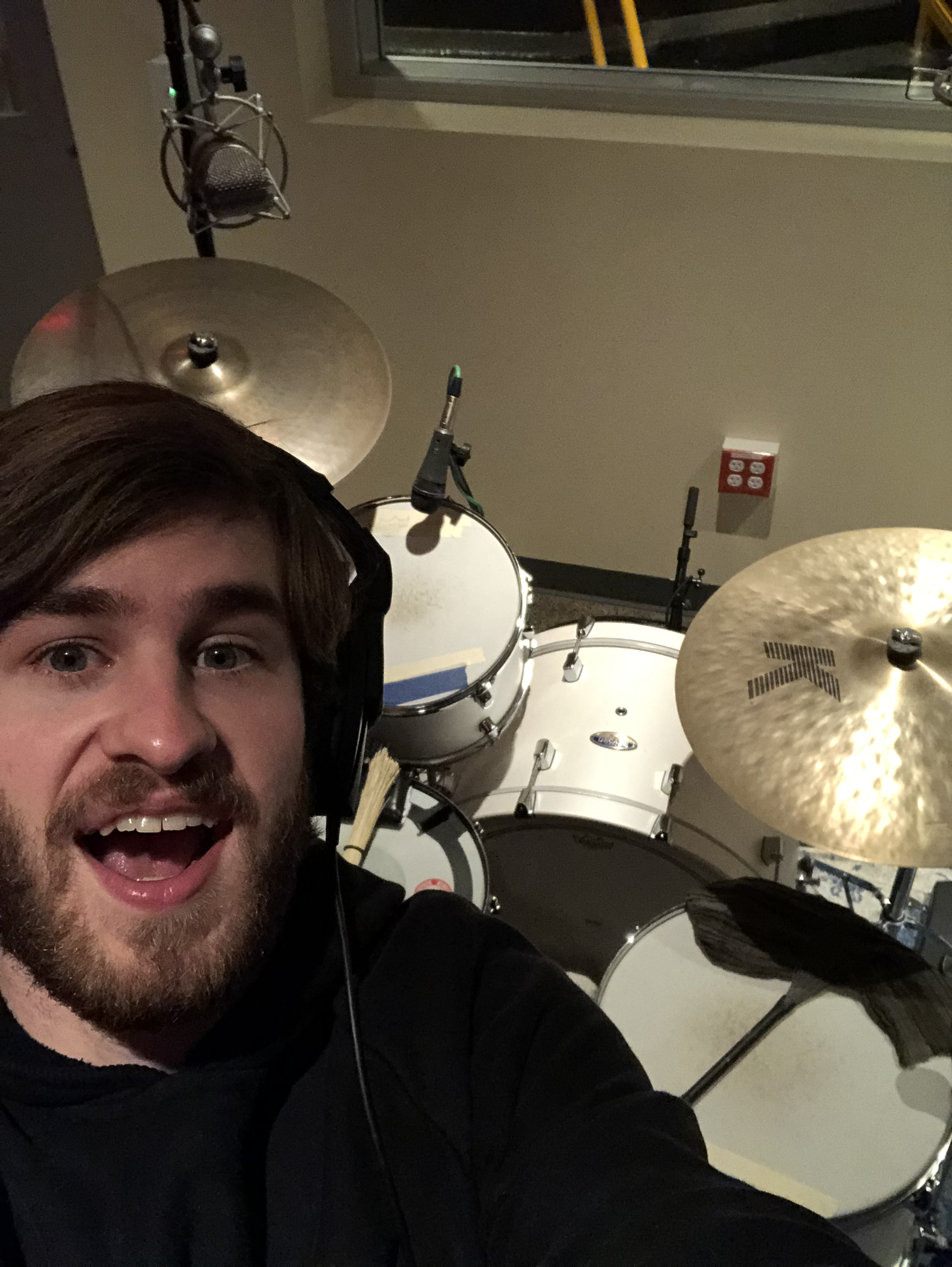 A man wearing headphones and a black hoodie taking a selfie in front of a drum set in a recording studio.