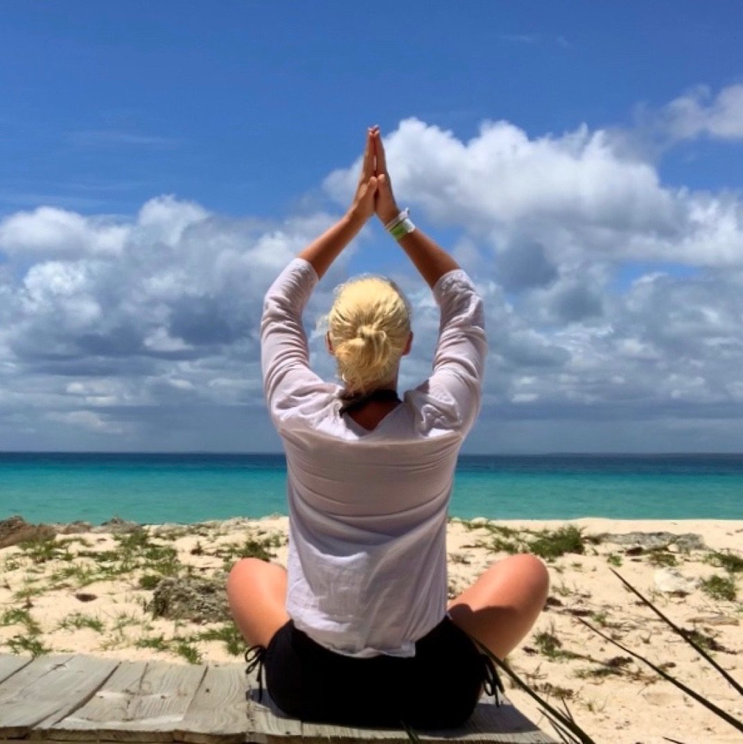 A woman with blonde hair tied in a bun practicing yoga in a seated meditation position on a wooden platform at the beach, facing the ocean with blue sky and clouds overhead.