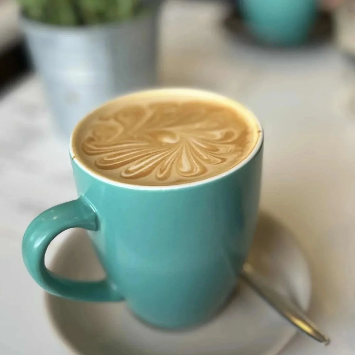 A teal mug filled with coffee featuring latte art in a floral pattern, placed on a white surface with a spoon beside it.