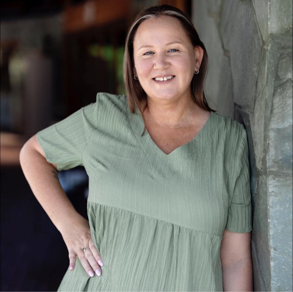 A woman with shoulder-length brown hair, smiling, standing next to a stone wall, wearing a light green dress.