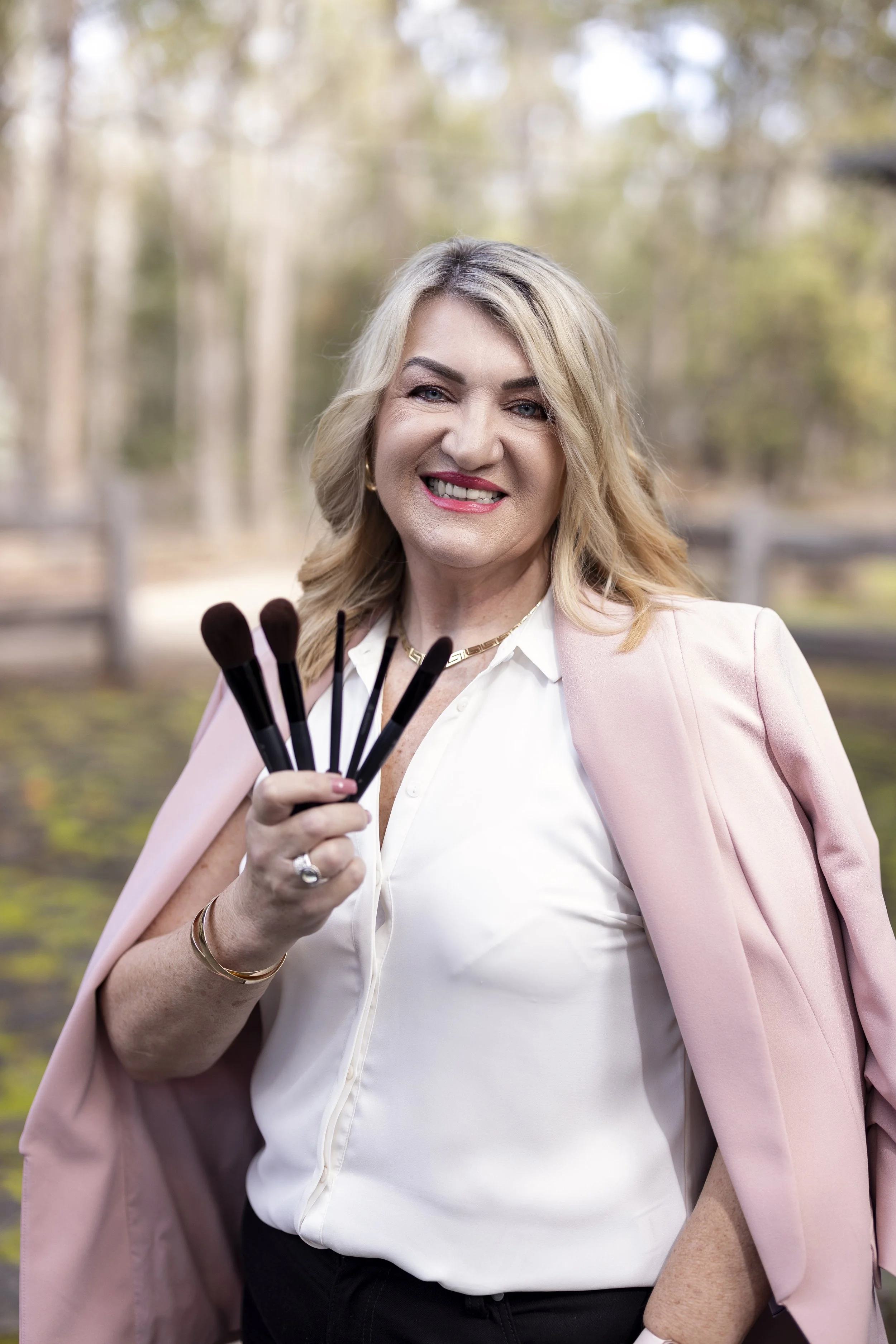 A woman smiling and holding five makeup brushes outdoors, dressed in a white blouse and pink blazer, with blurred trees in the background.