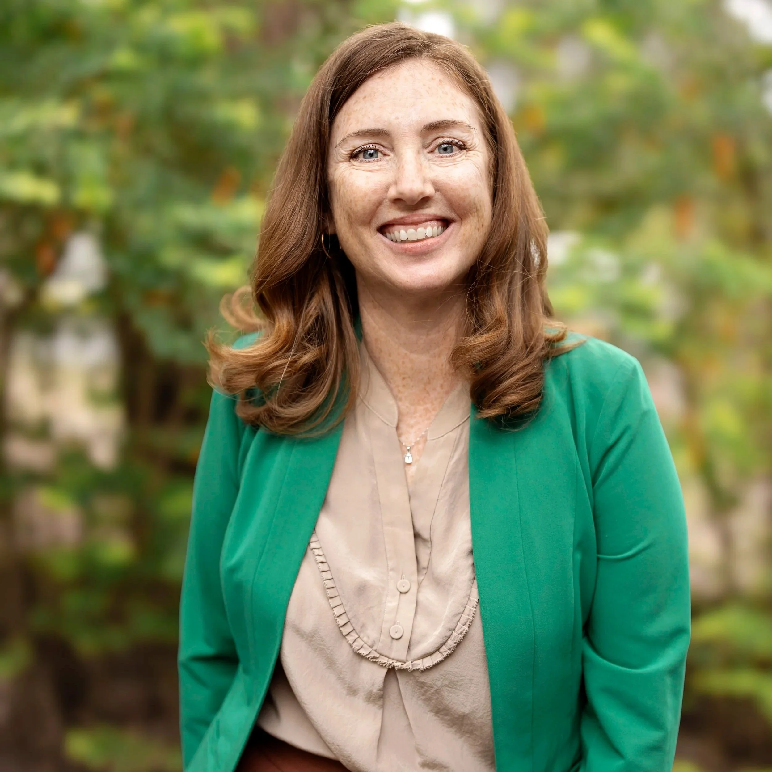 A woman with red hair, blue eyes, and freckles smiling outdoors in front of green foliage, wearing a green blazer and beige blouse.