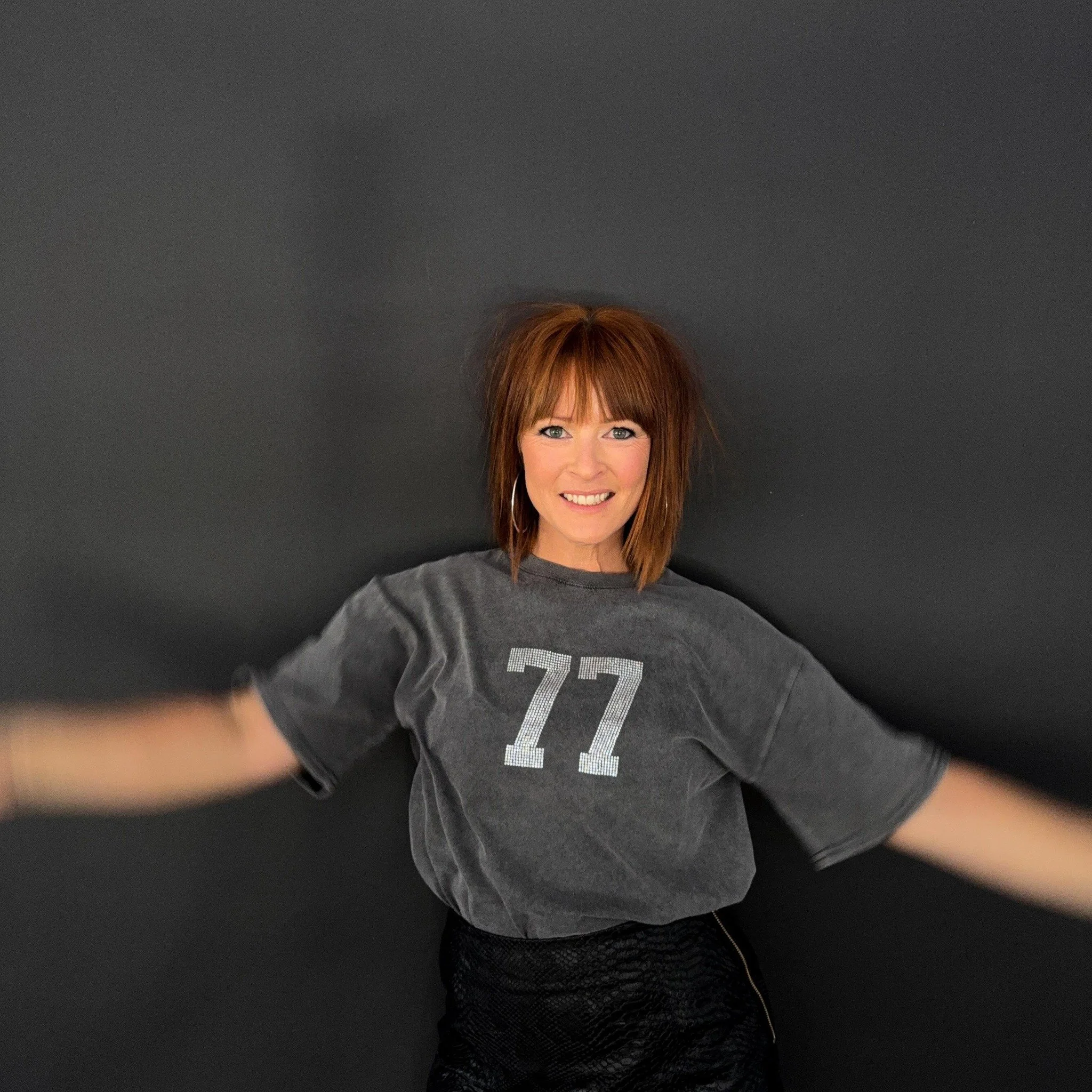 A woman with red hair and a bright smile, wearing a gray t-shirt with the number 77 on it, standing against a black background.