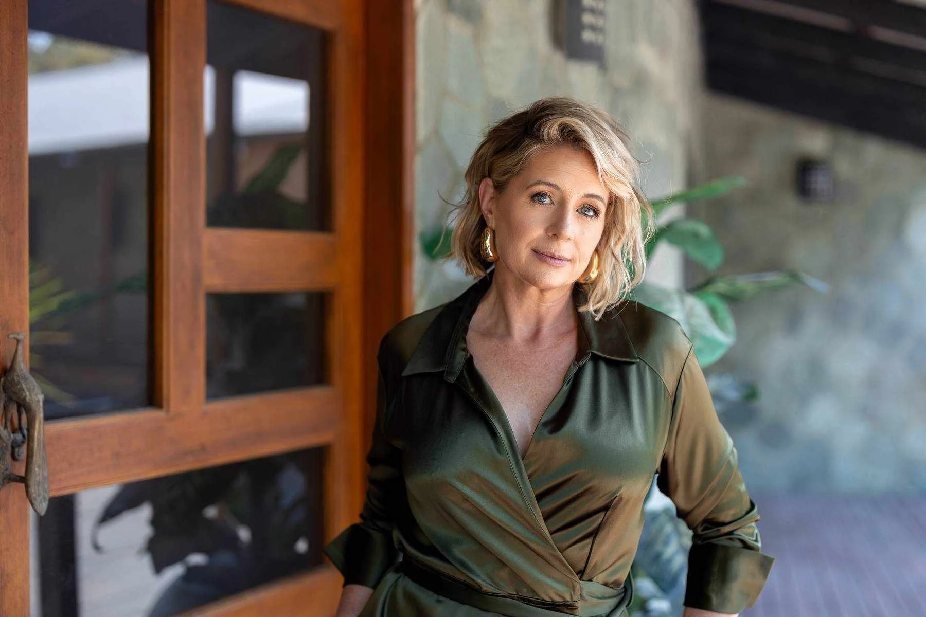 A woman with shoulder-length blonde hair wearing a dark green satin blouse with rolled sleeves, standing in front of a wooden door with glass panels and a stone wall, looking at the camera.