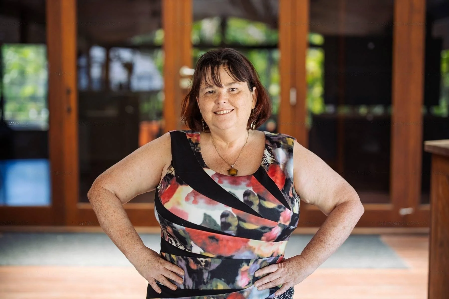 A woman with short, dark brown hair and freckles, smiling, standing indoors with hands on her hips. She is wearing a sleeveless dress with a colorful floral pattern and a necklace with a rectangular pendant.