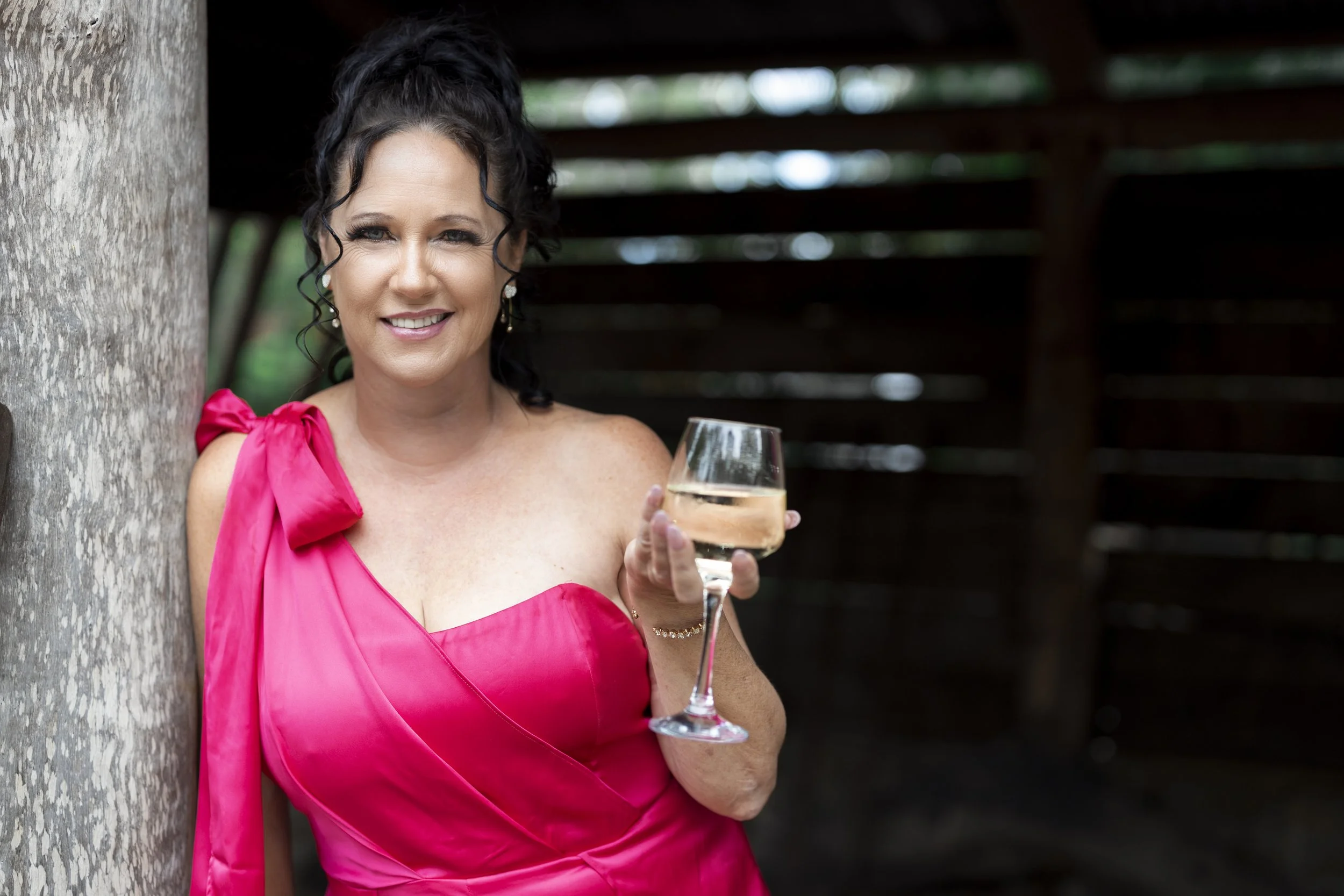 A woman in a bright pink dress holding a glass of white wine, standing outdoors near a tree, smiling at the camera.