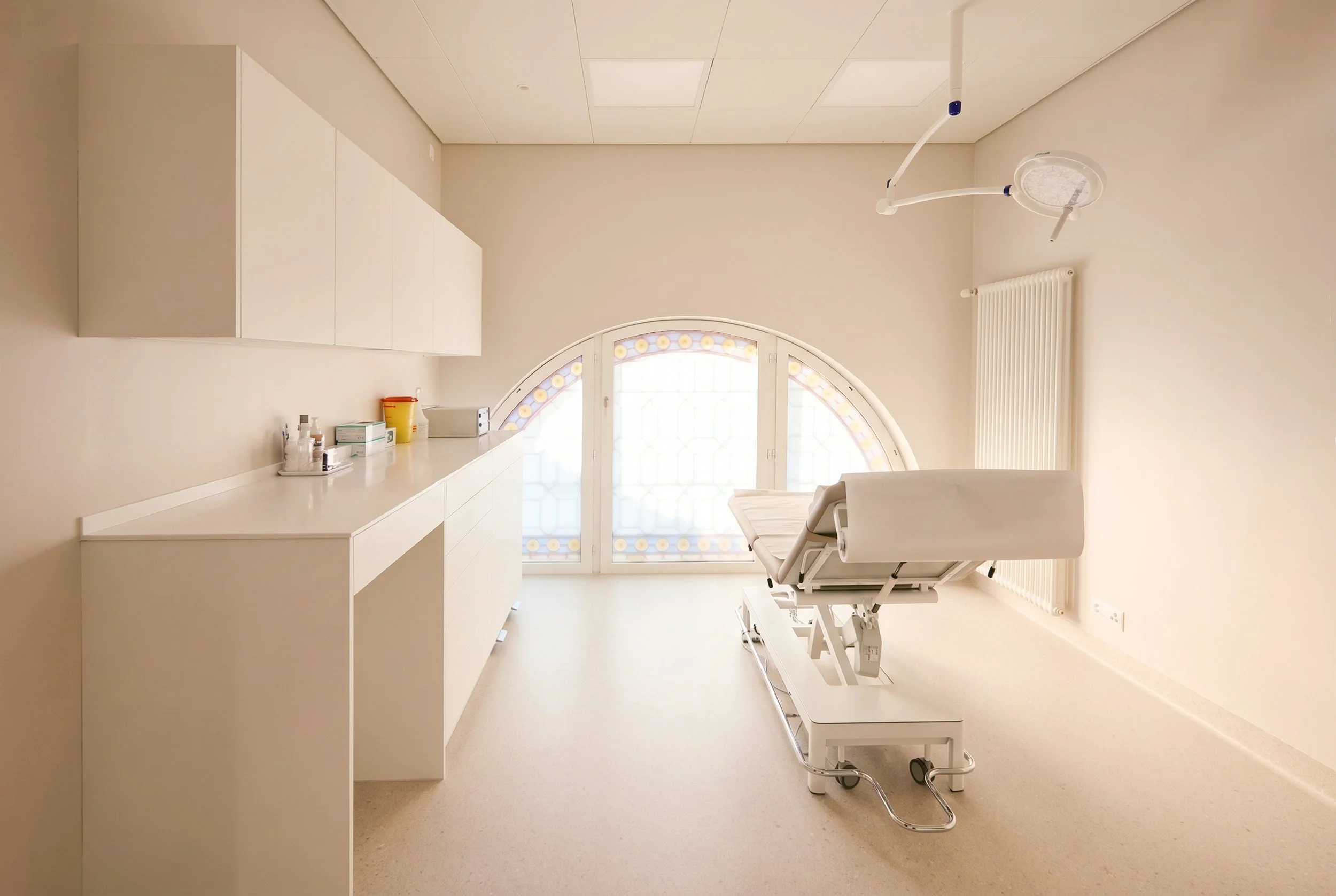 Empty medical examination room with a white adjustable examination table, a white chair, a tall floor lamp, a window, and wood flooring.