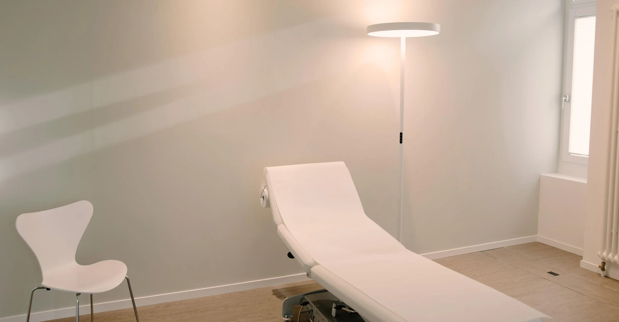 Empty medical examination room with a white adjustable examination table, a white chair, a tall floor lamp, a window, and wood flooring.