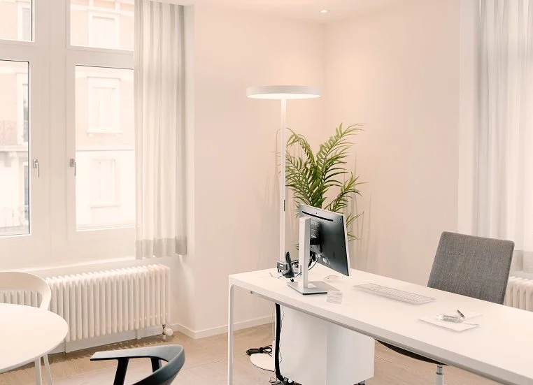 Modern office space with a white desk, desktop computer, gray office chair, large window with white curtains, potted plant, and a tall standing lamp.