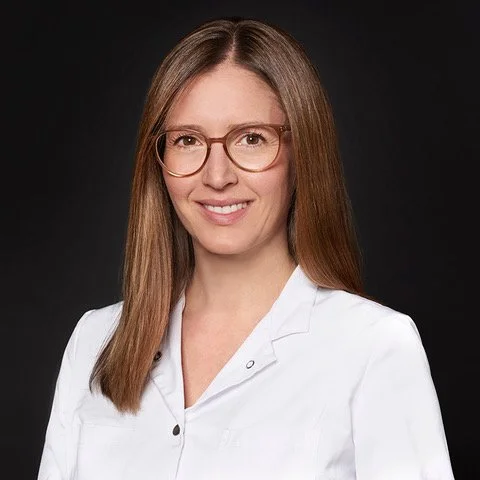 A woman with long, straight hair wearing glasses and a white medical coat, smiling against a dark background.