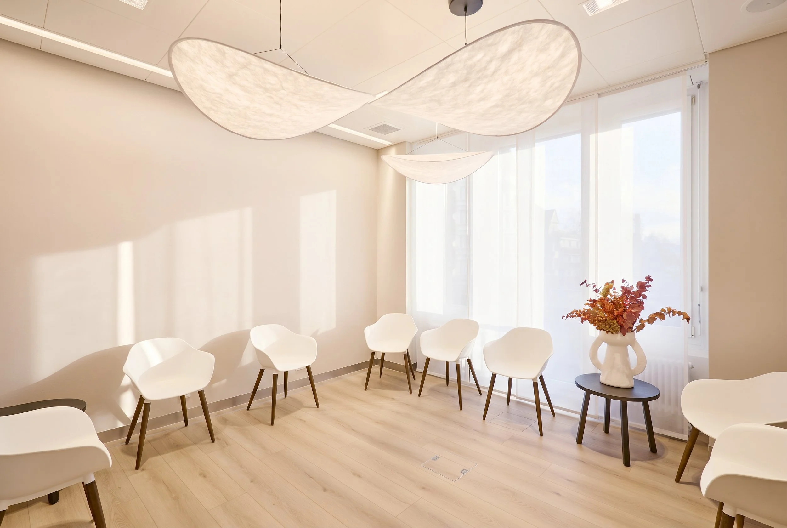 A minimalist interior with a dark brown bottle diffuser and reed sticks on a white floating shelf, a wooden slat wall behind, and a white chair with a black desk or table with a keyboard in the background.
