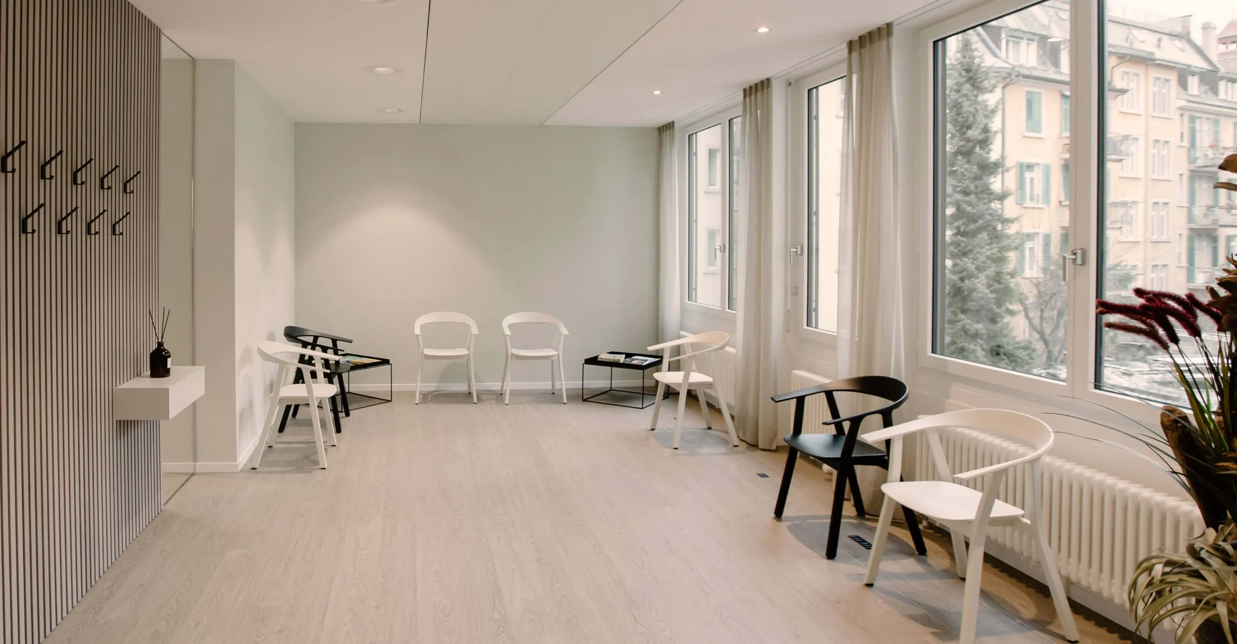 Modern waiting room with white and black chairs, large windows with beige curtains, light wooden floors, and a minimalist decor including a small wall shelf with a vase and reed diffuser.