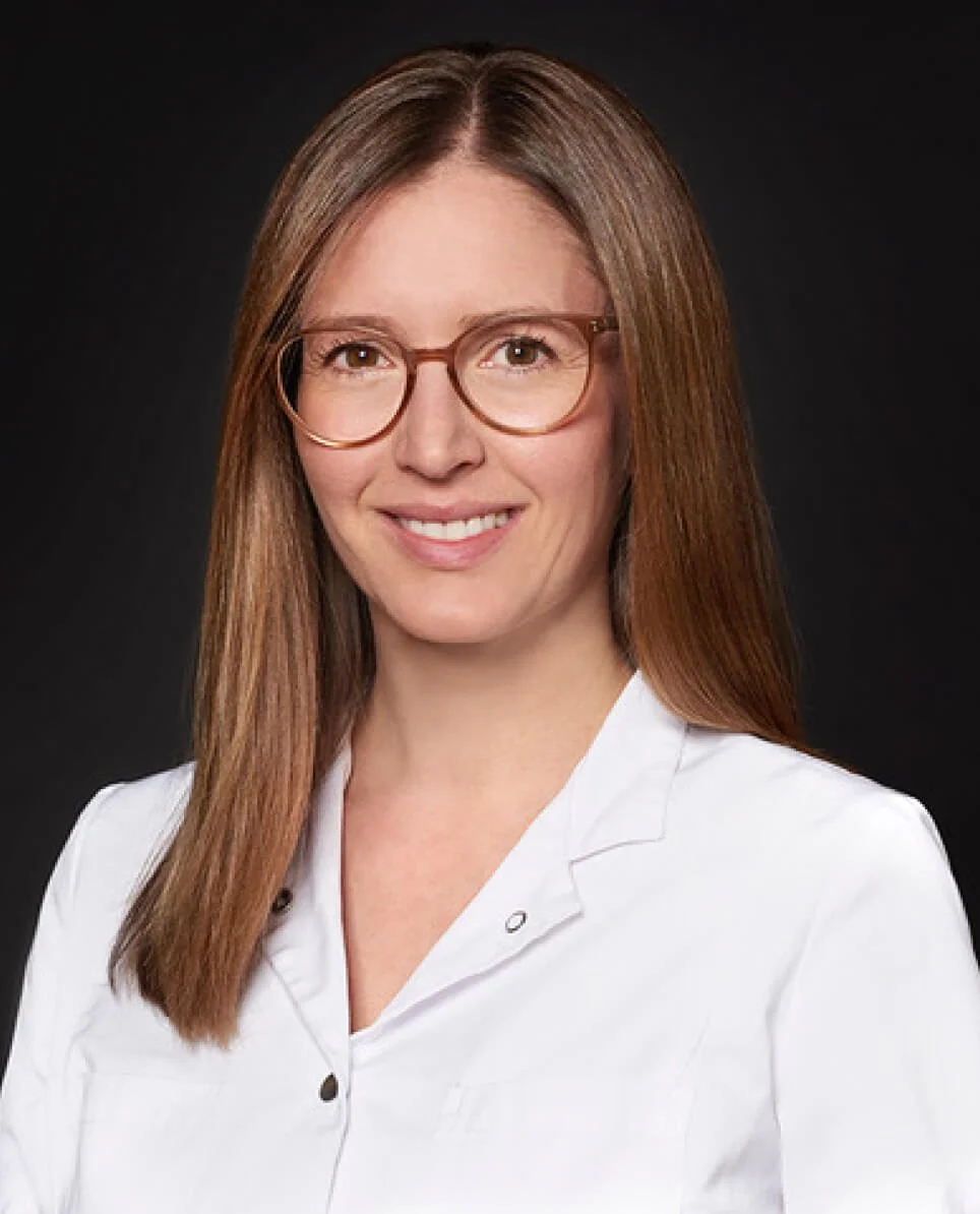 A woman with shoulder-length brown hair, wearing glasses and a white lab coat, smiling against a dark background.