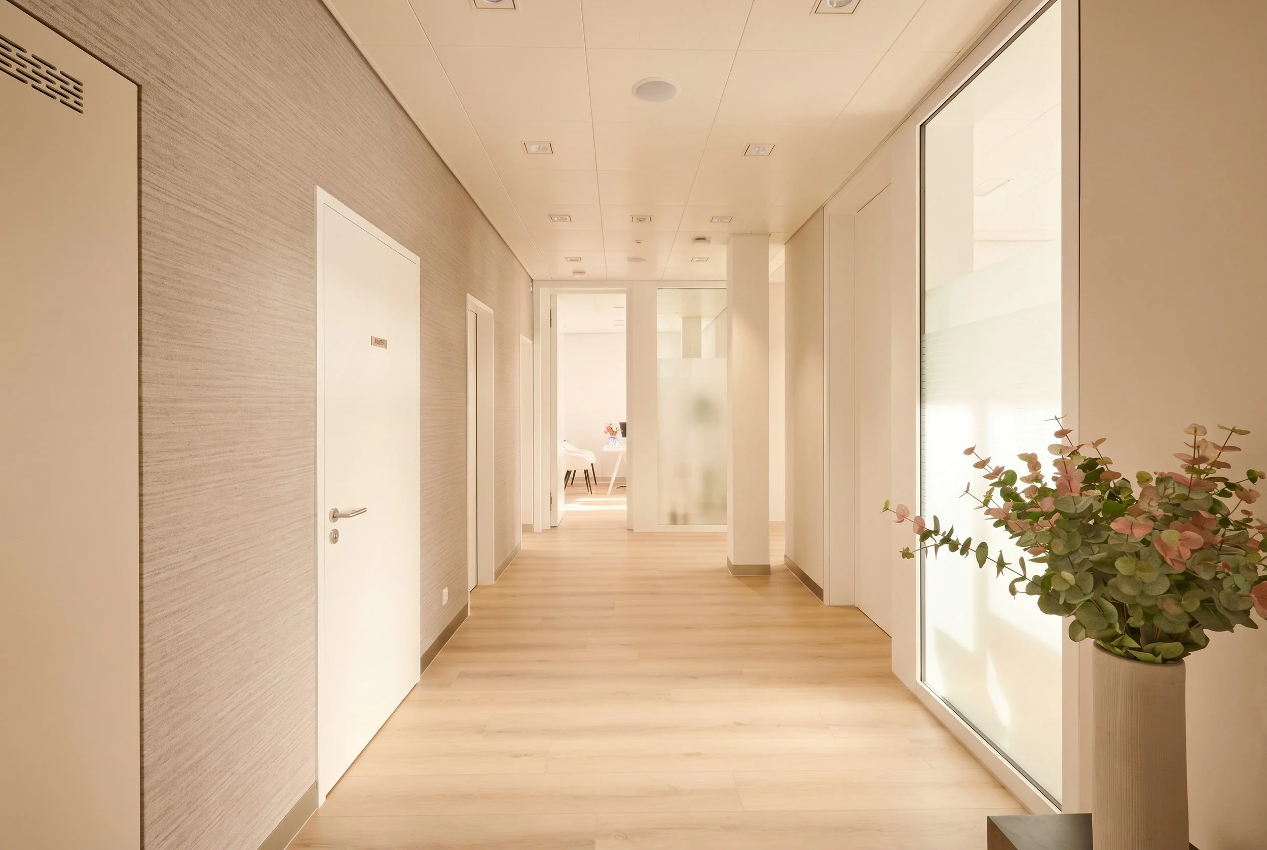 Modern waiting room with white and black chairs, large windows with beige curtains, light wooden floors, and a minimalist decor including a small wall shelf with a vase and reed diffuser.