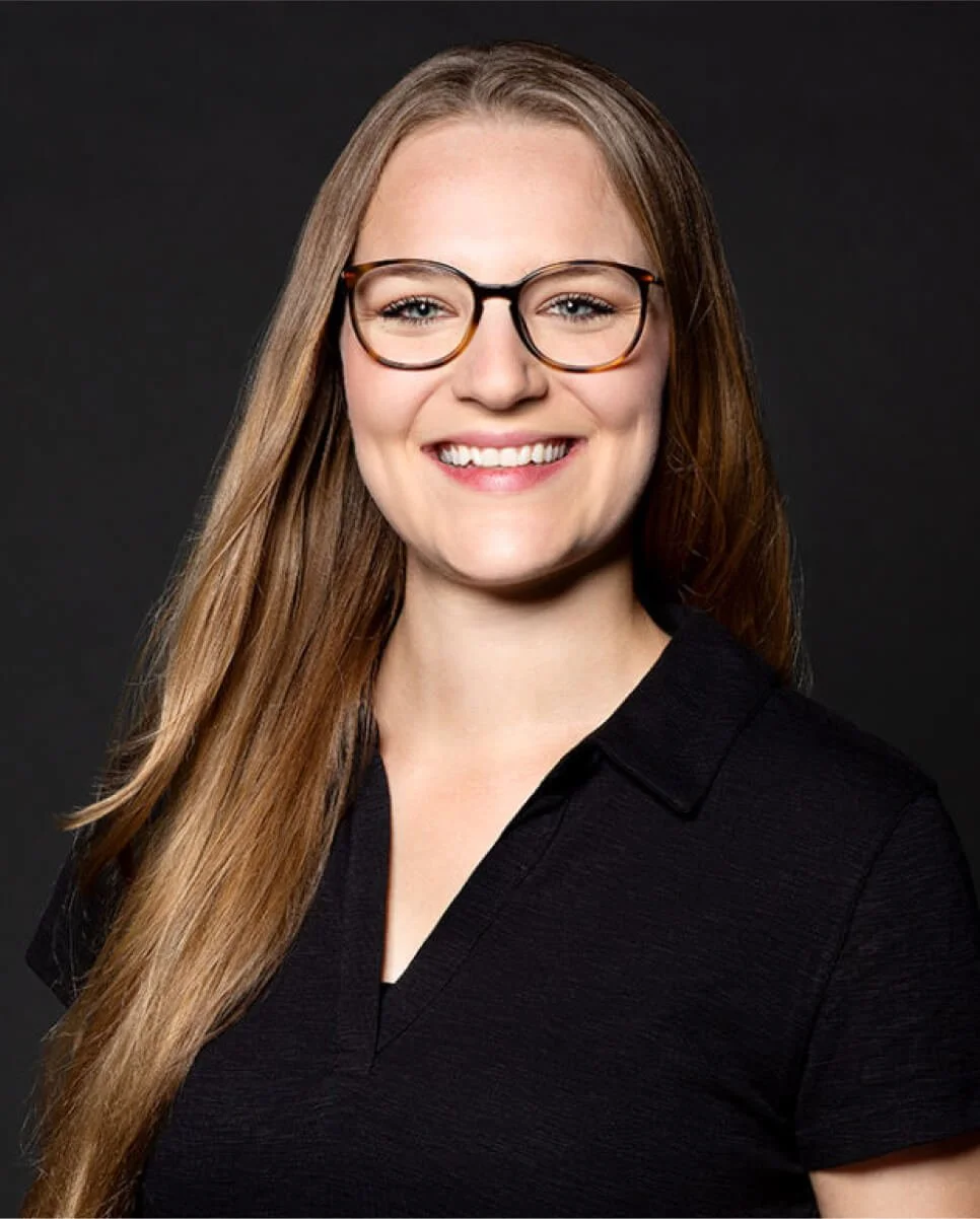 A young woman with long, straight, light brown hair, wearing black-rimmed glasses and a black collared shirt, smiling against a dark background.