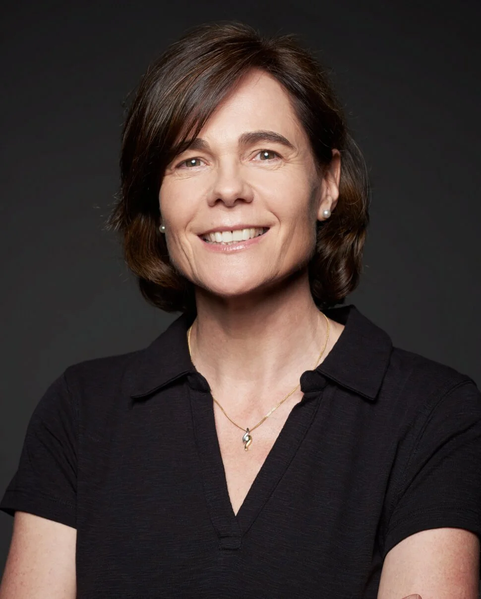 Portrait of a smiling woman with short brown hair wearing a black collared shirt, pearl earrings, and a gold necklace with a swirl pendant, against a dark background.