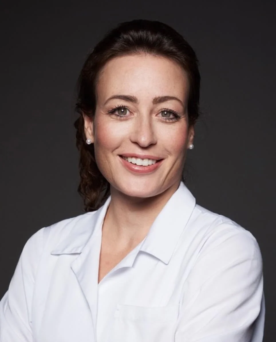 Professional woman with brown hair, wearing pearl earrings and a white collared shirt, smiling against a dark grey background.