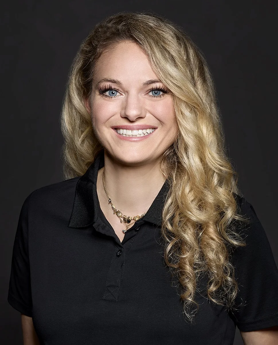 Portrait of a smiling woman with blue eyes, blonde curly hair, wearing a black collared shirt and a gold necklace, against a dark background.