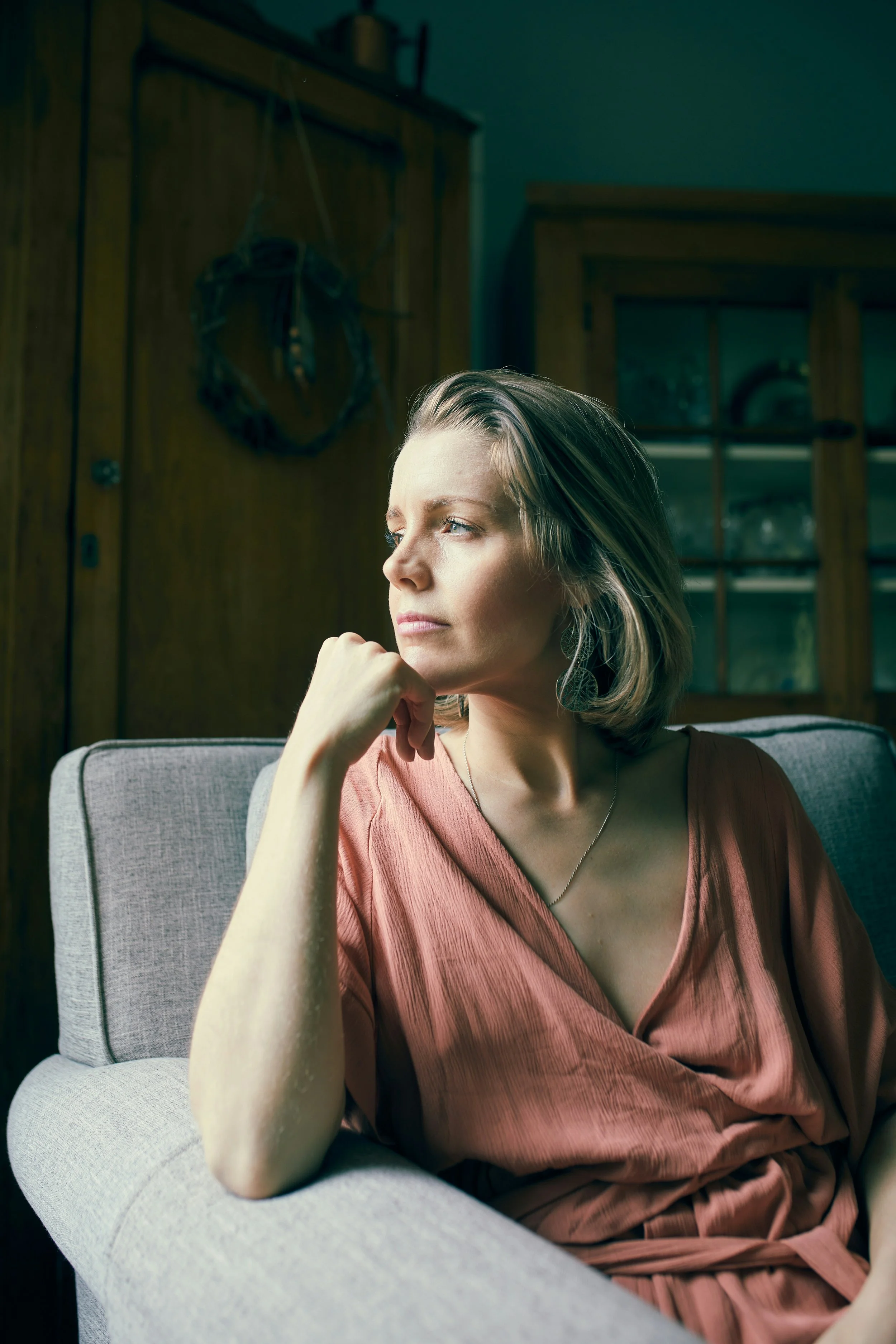 A woman with blonde hair wearing a pink dress sitting on a light gray sofa in a room with wooden cabinets and a dark teal wall, looking thoughtfully to the side - individual therapy for identity and codependency.