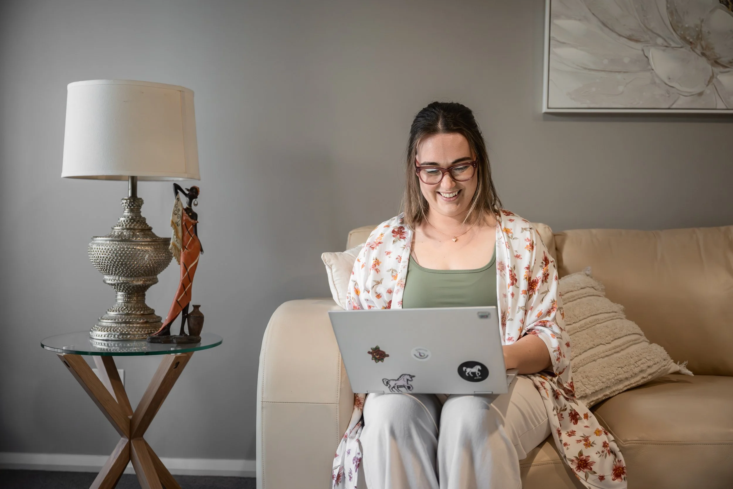 A woman wearing glasses and a floral cardigan sitting on a beige couch, working on a laptop with stickers, in a cozy living room with a lamp and artwork on the wall.