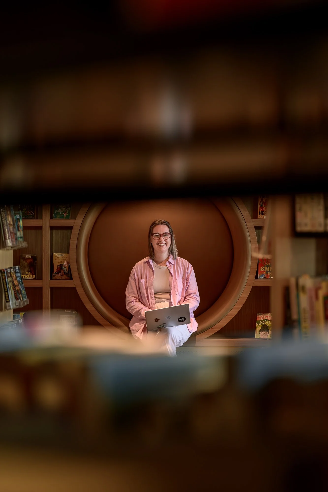 Young woman with glasses smiling and holding a laptop, seated on a wooden circular nook in a library, surrounded by bookshelves with books.