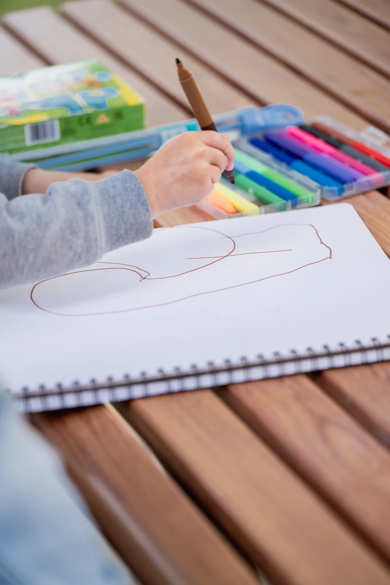 Child drawing with a brown marker on a white sketchbook on a wooden table, with colored markers and a box of art supplies nearby.