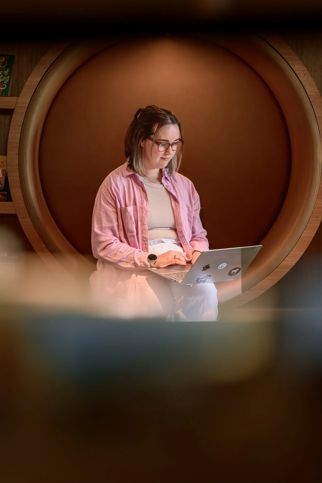 A woman sitting inside a circular wooden structure, working on a laptop with stickers on it, in a cozy, dimly lit room.