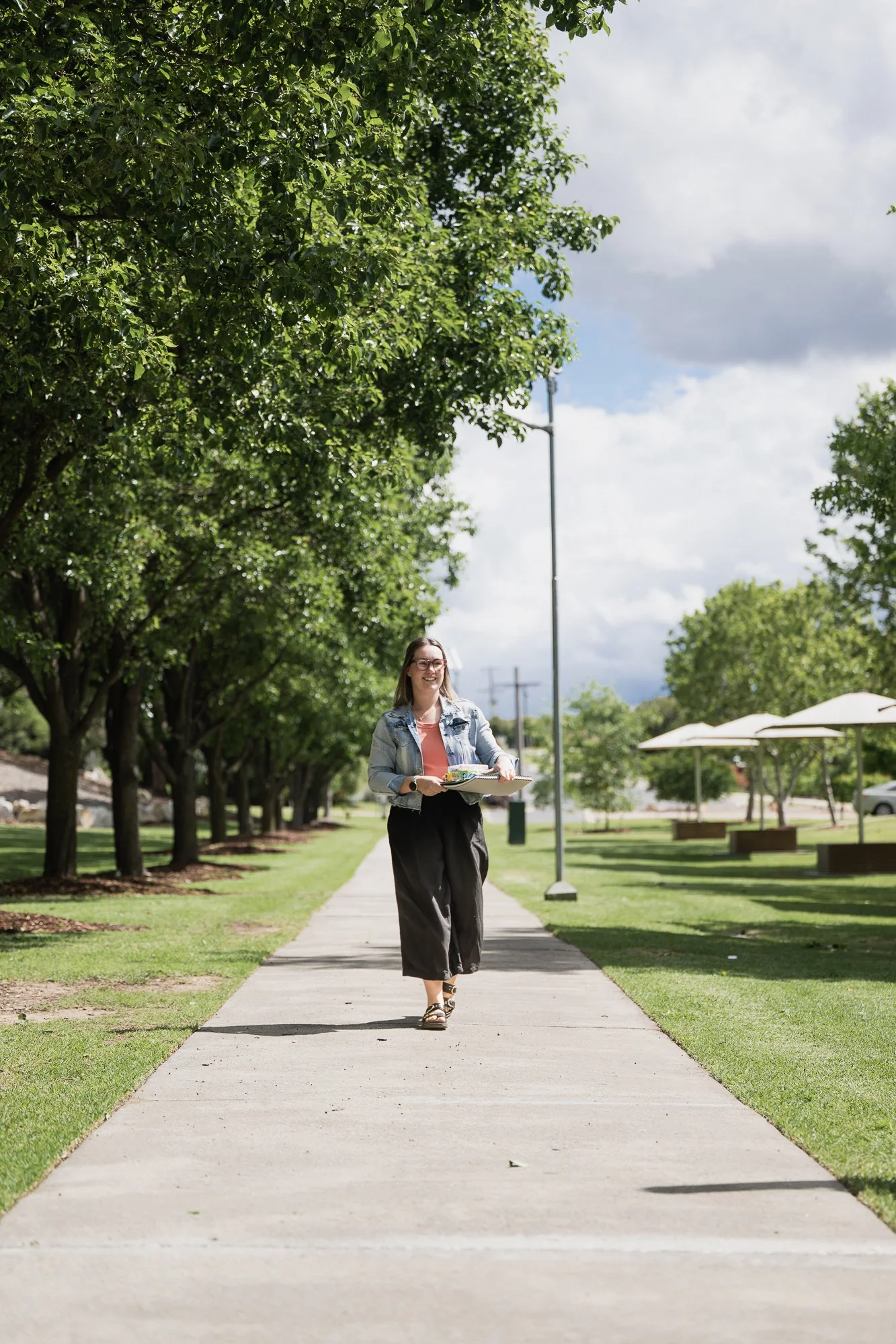 A woman with glasses and a denim jacket walking on a sidewalk in a park, holding a tray with food, trees lining the path, and umbrellas in the background.