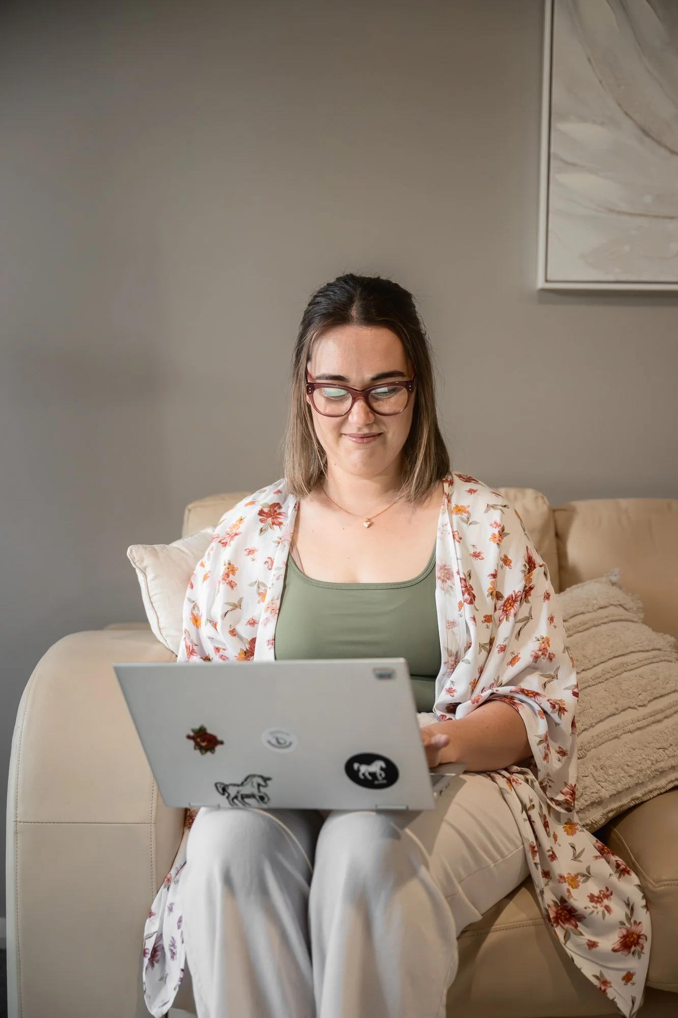 A woman with glasses sitting on a beige couch, using a laptop decorated with stickers, in a living room with neutral-colored walls and artwork hanging on the wall.