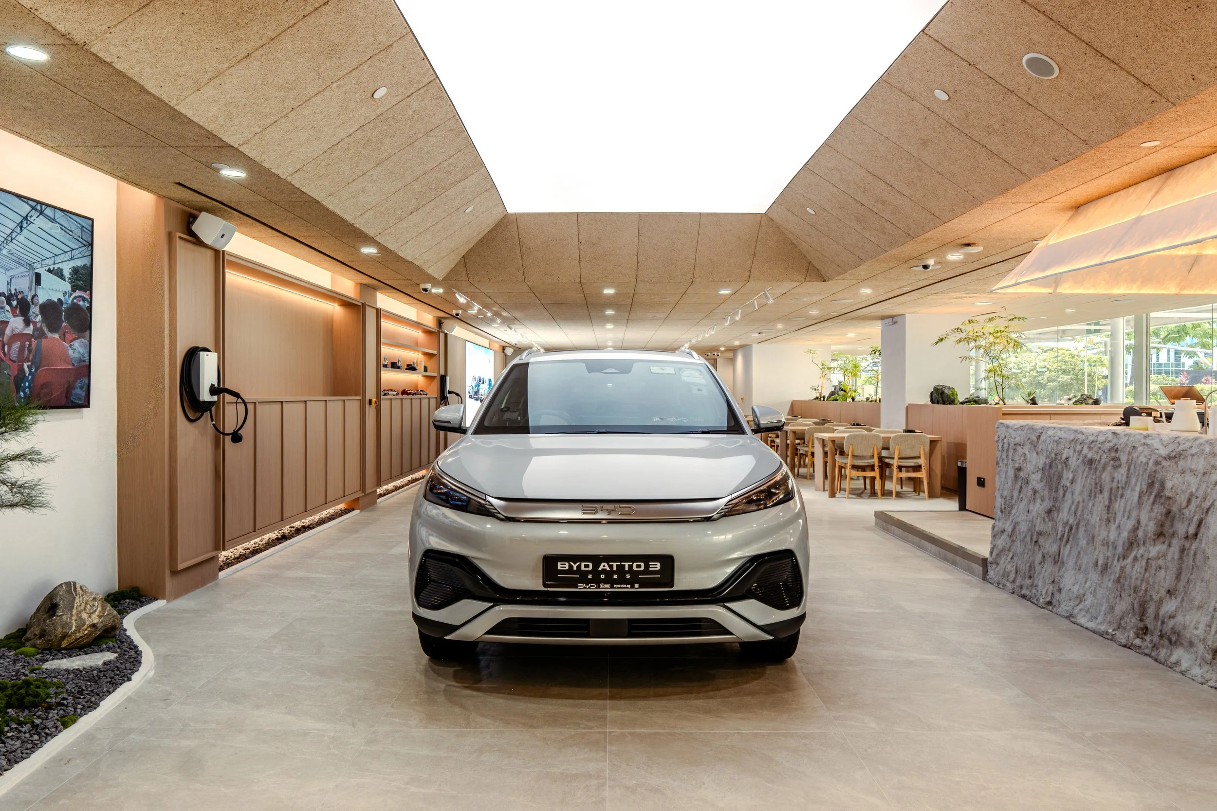 A white BYD ATTO 3 electric vehicle parked indoors in a modern showroom with wooden and glass decor, seating area, and indoor plants.
