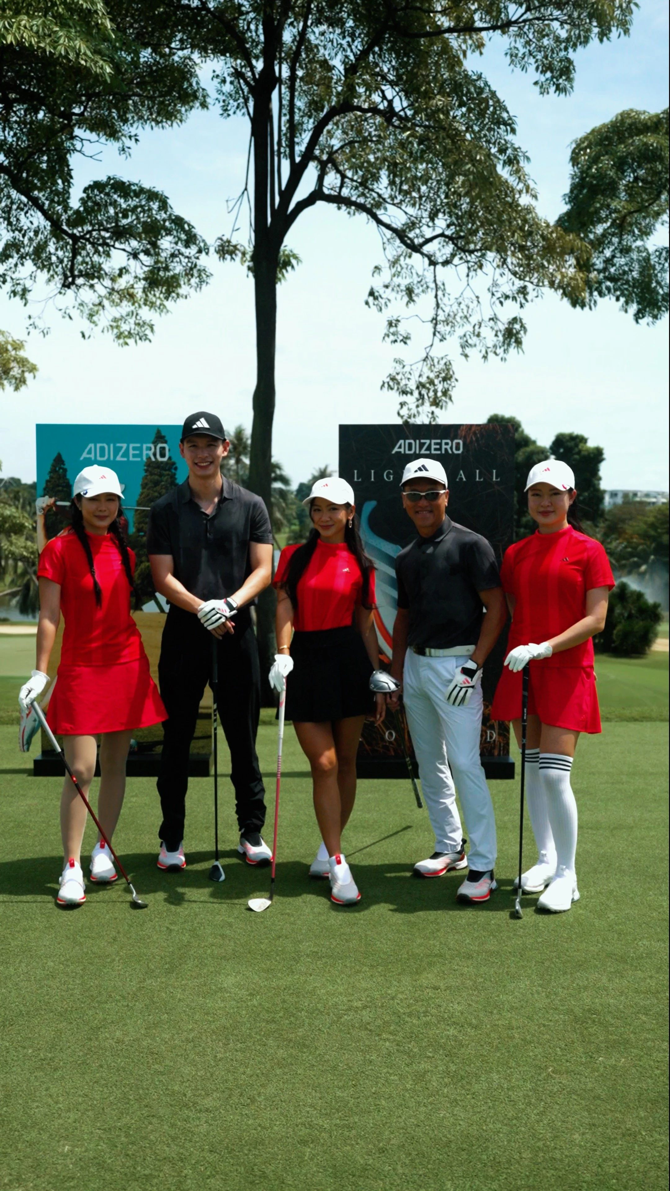 Group of five people on a golf course, posing with golf clubs, four women wearing red dresses and white hats, one man wearing black shirt and white pants, with banners in the background.