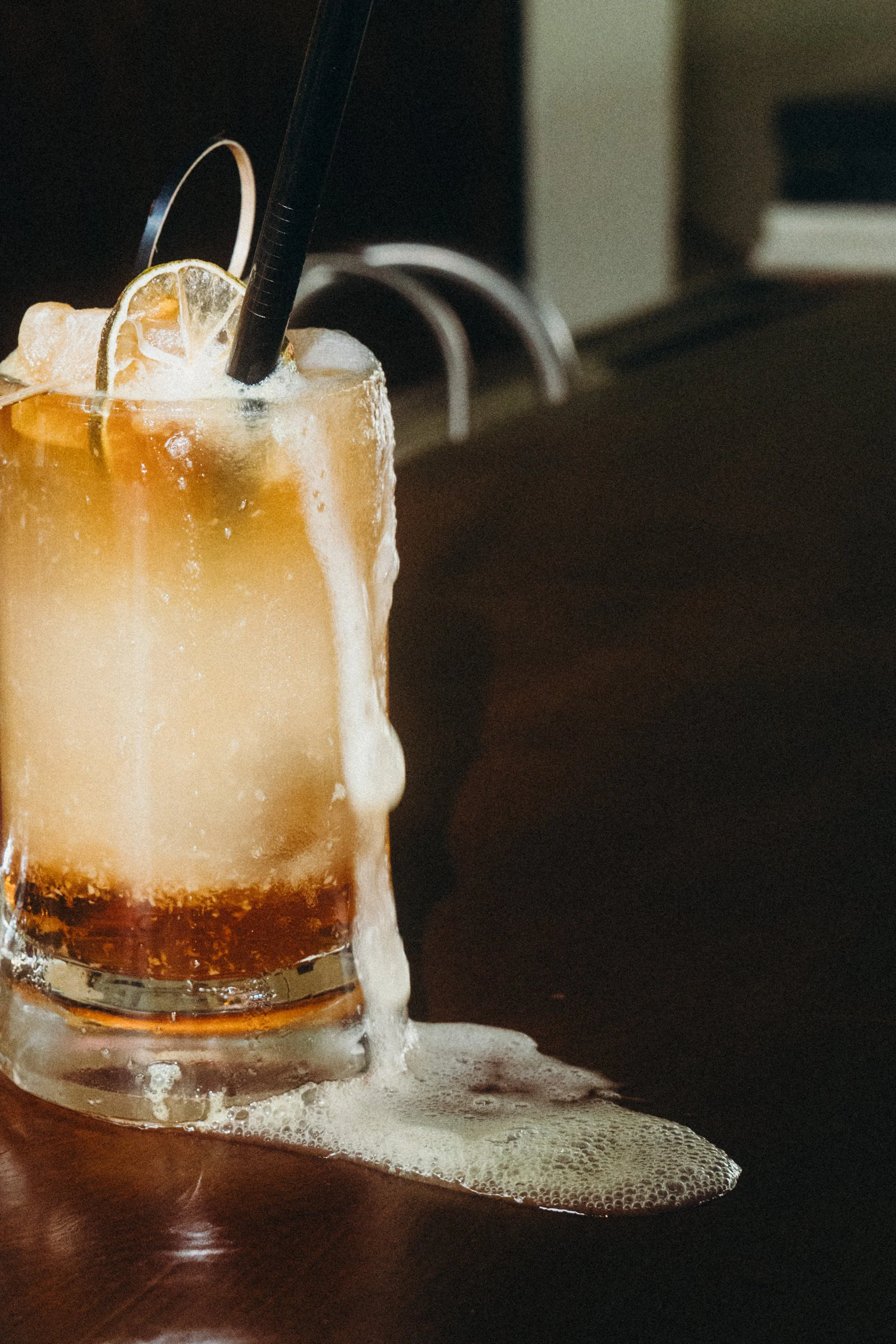 Mine 'n' Stormy, house cocktail with home made ginger beer and lemon slices and a black straw, overflowing and spilling foam onto a dark wooden surface, the bar