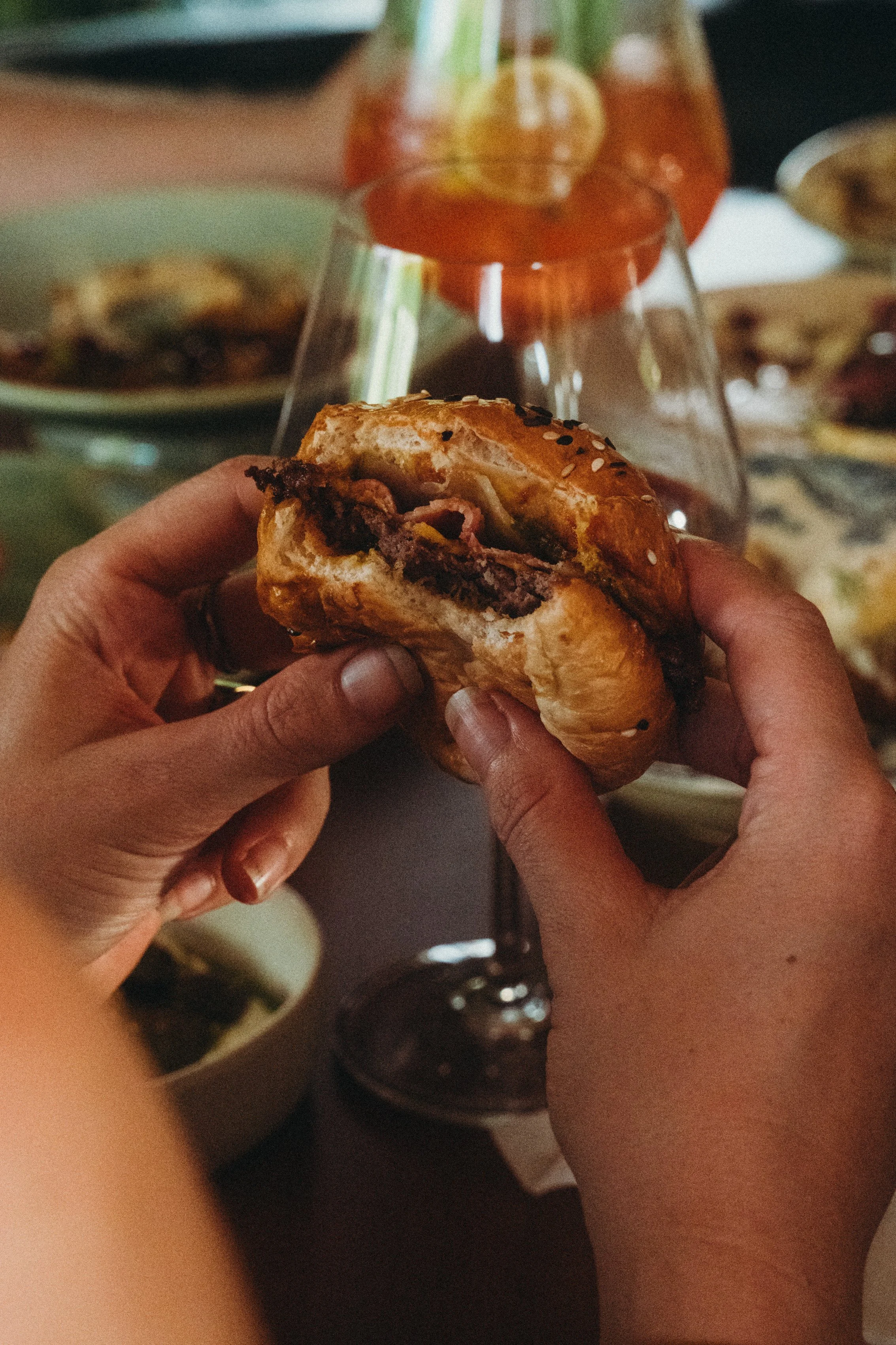 Girl holding a half-eaten burger with a toasted bun and visible beef patty with an Aperol cocktail in the background on a table with other dishes of The Lakehouse
