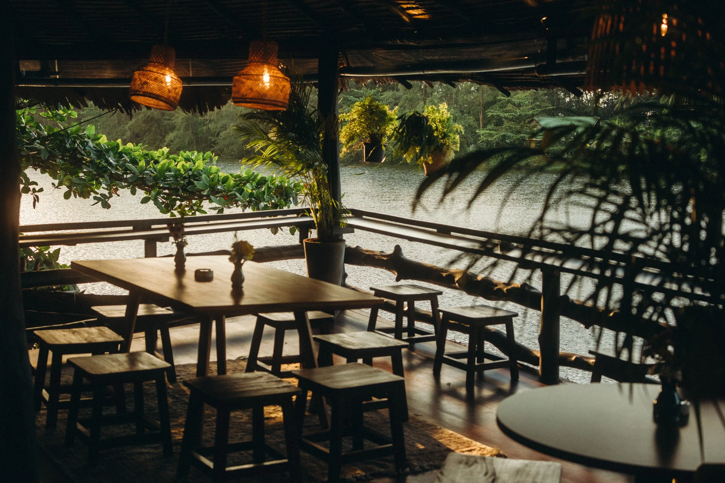 A rustic outdoor restaurant with wooden tables and stools by a body of water, decorated with hanging wicker lights and potted plants, during sunset.