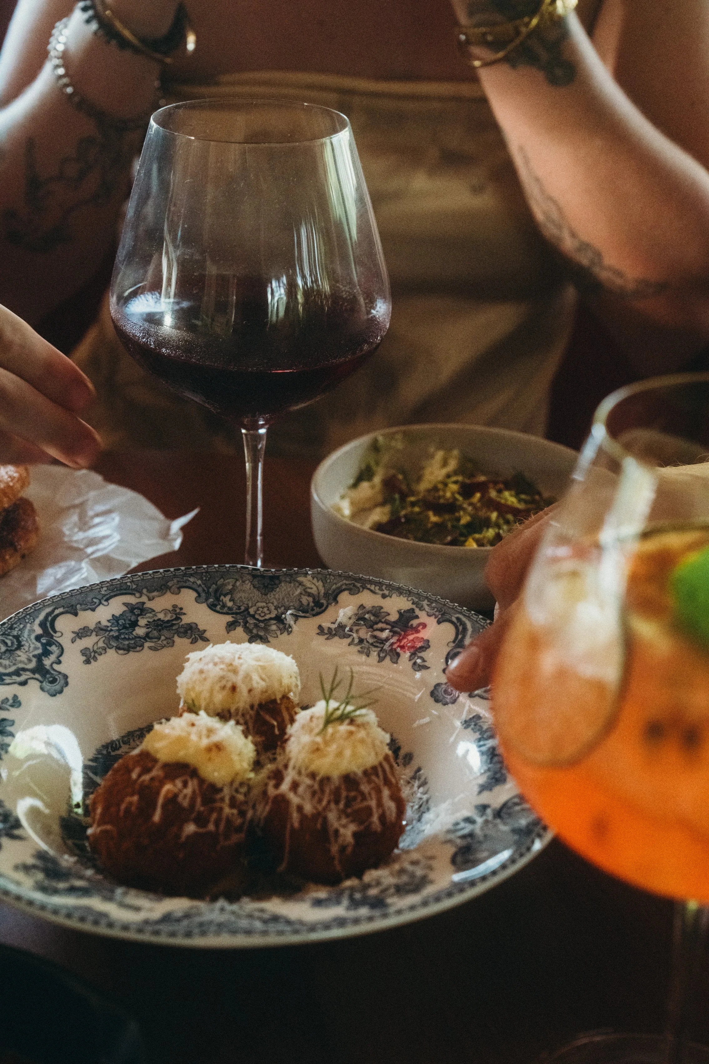 Table full of food with people enjoying a meal with a glass of red wine, stracciatella dip, chorizo arancinis with parmesan cheese, and a colorful aperol cocktail